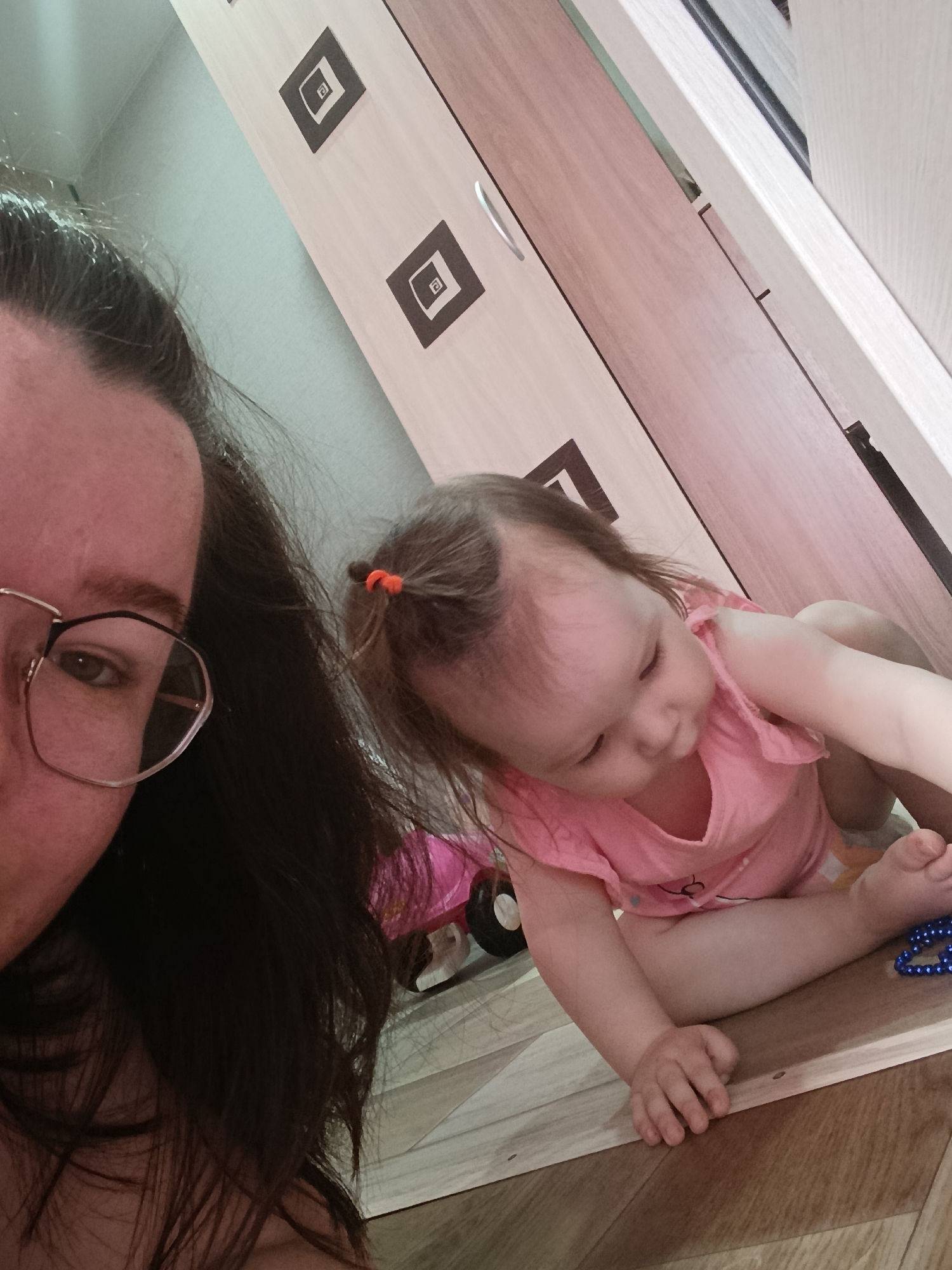 An indoor, close-up selfie captures a moment between a woman and a baby, likely taken in a home in Chaikovsky, Russia. The woman's face is partially visible on the left side of the frame, showing her left eye and forehead, framed by dark hair and rectangular glasses with a two-tone frame. Her expression appears neutral.
To the right, a baby, appearing to be a toddler, is on a light wood-patterned laminate floor. The baby is wearing a pink top and has light brown hair tied up with a small orange elastic band on top of their head. They are looking downwards, with their hands resting on the floor. Near the baby's right hand, a blue beaded item, possibly a necklace or toy, is visible. Part of a pink toy with wheels is also visible behind the baby on the left.
In the background, a light-colored wooden wardrobe with dark square patterns is prominent. Each dark square contains nested smaller squares, with the innermost small square featuring a faint 'a'-like symbol. The scene suggests a casual, domestic setting during daytime.