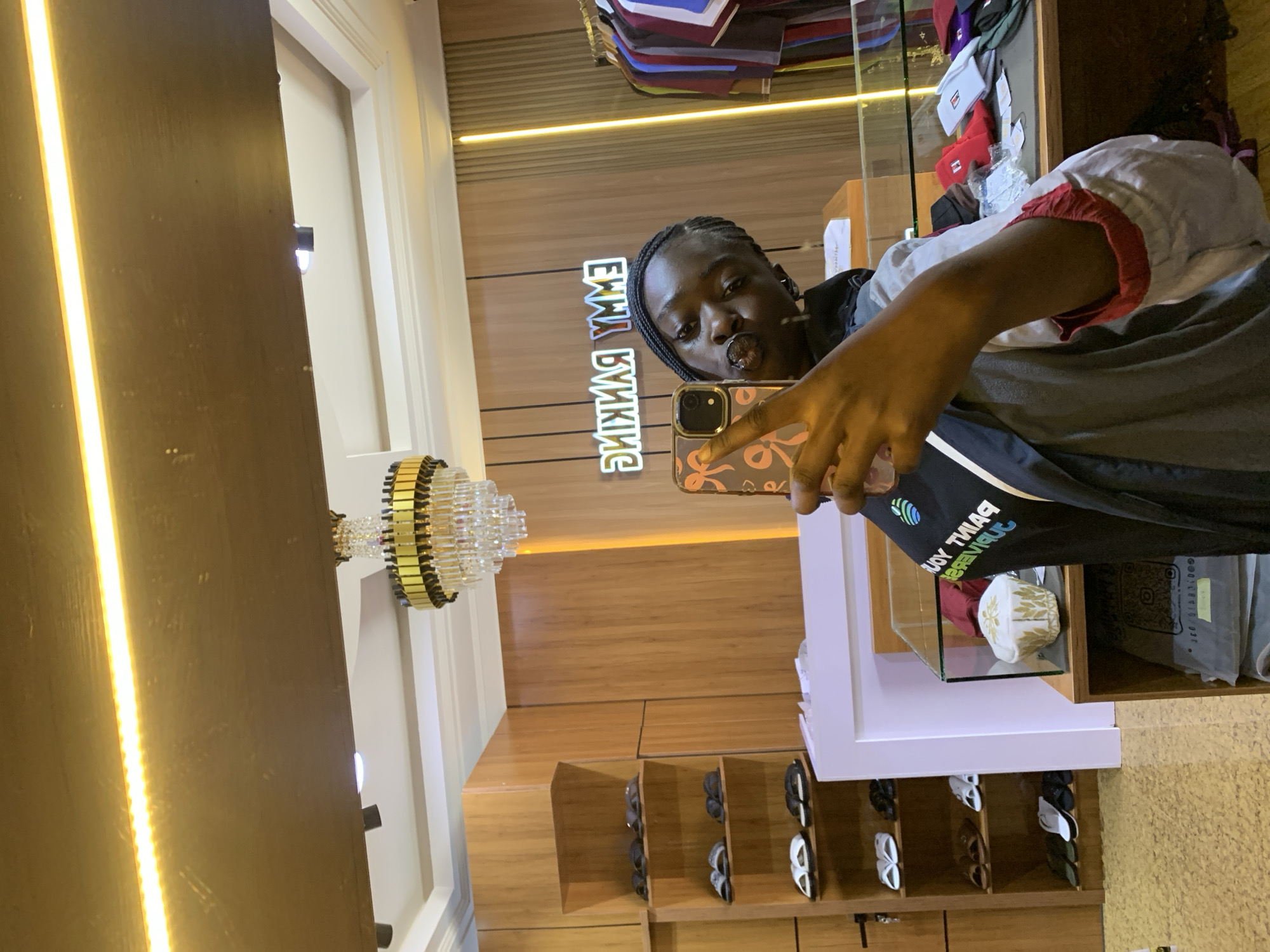 The image depicts a young woman taking a mirror selfie in a retail environment that appears to be a clothing or shoe store, potentially located in Gura Top, Nigeria. The store features wooden paneled walls and shelving units displaying footwear. A prominent neon sign with reversed lettering, reading "EMMY RANKING," is visible on the wall. The woman is wearing a dark jacket with a white and red trim, and a black garment underneath. She is holding a smartphone with a patterned case in her right hand and has her left hand raised, with her index and middle fingers extended. A chandelier hangs from the ceiling, and display cases containing hats and other merchandise are present. The lighting is bright, with an LED strip illuminating the ceiling area.