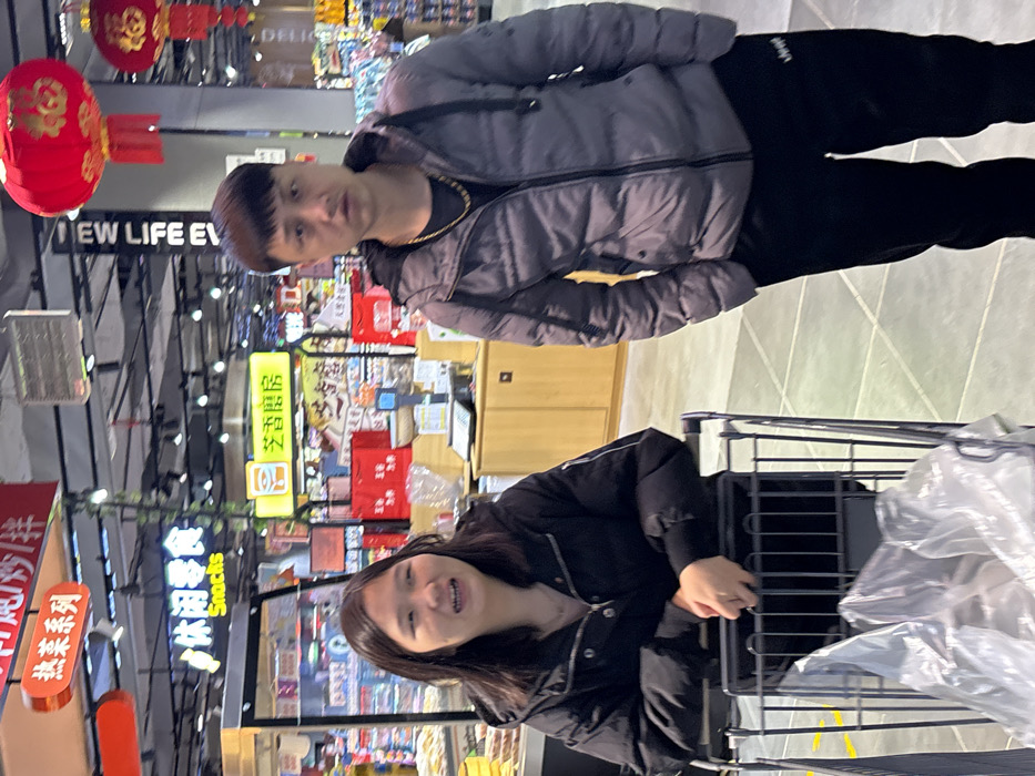 This image depicts an indoor scene within a supermarket or hypermarket in Jiangjin, China. The setting features a light-colored tiled floor and overhead lighting. Two individuals are present in the foreground. On the left, a female subject with dark, shoulder-length hair is partially visible, wearing a black puffer jacket over a light top. She holds the handle of a black metal shopping cart, which contains a clear plastic bag. Her mouth is slightly open, and she appears to be speaking or smiling. On the right, a male subject with short, dark hair stands facing the camera. He wears a grey puffer jacket, a dark t-shirt with a gold-colored chain, and black track pants featuring a white 'Jeep' logo on the left thigh. His hands are in his pockets. In the background, various illuminated signs display Chinese characters, including "热菜系列" (Hot Dish Series), "休闲零食 Snacks" (Leisure Snacks), and "芝香居易" (Zhixiang Juyi). Red Chinese lanterns with the character '福' (Fú - good fortune) are suspended from the ceiling. Store shelves, display cases, and a checkout counter with scales are visible in the mid-ground.