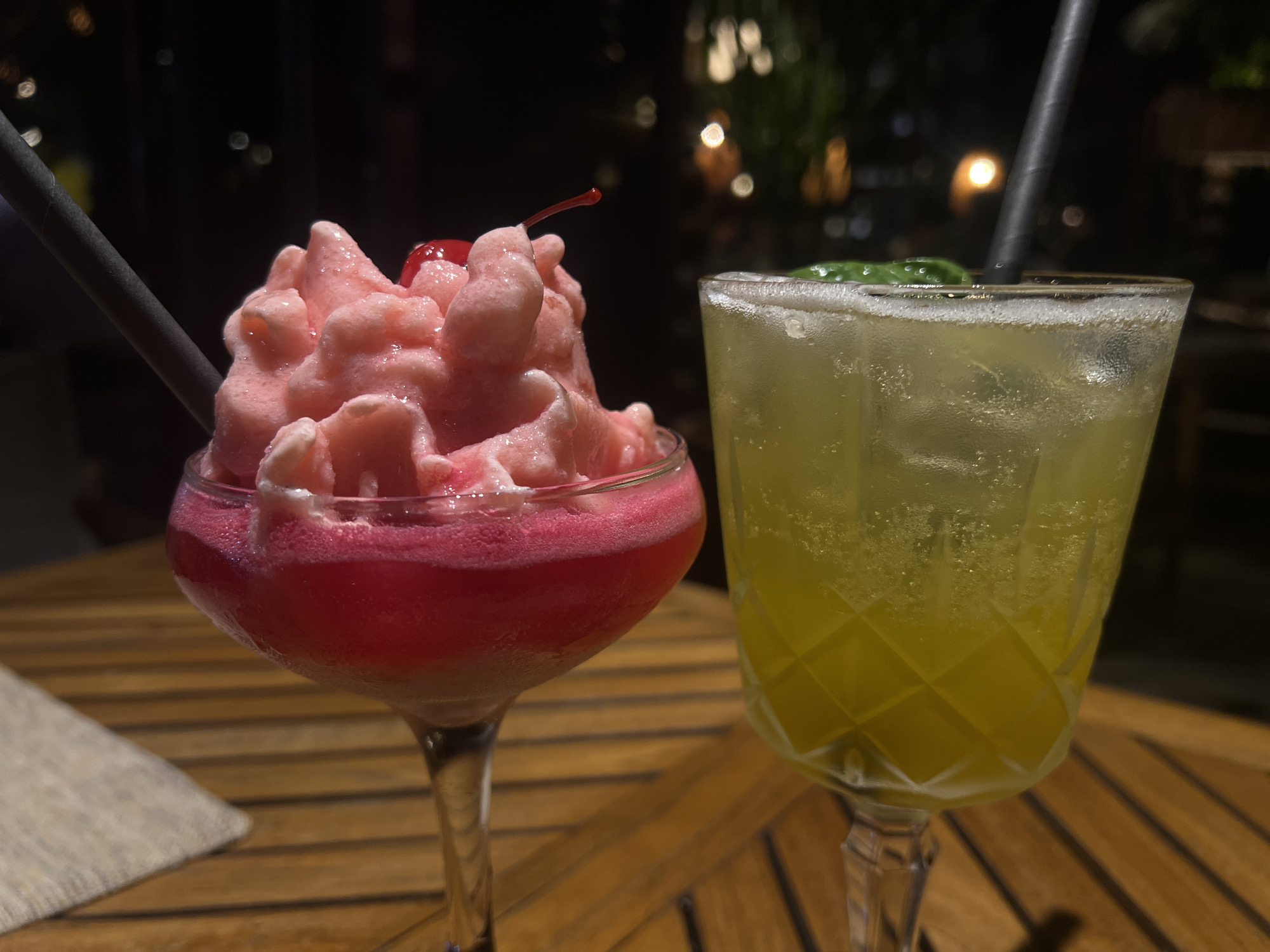 Two drinks are on a wooden table in an outdoor setting, likely in North Kuta, Indonesia. The drink on the left is a pink and red frozen beverage in a stemmed glass, topped with a cherry and a black straw. The drink on the right is a pale yellow liquid in a stemmed, cut-glass style glass, also with a black straw and a small green garnish. The table has a horizontal wooden slat pattern. The background is dark with blurred lights.
