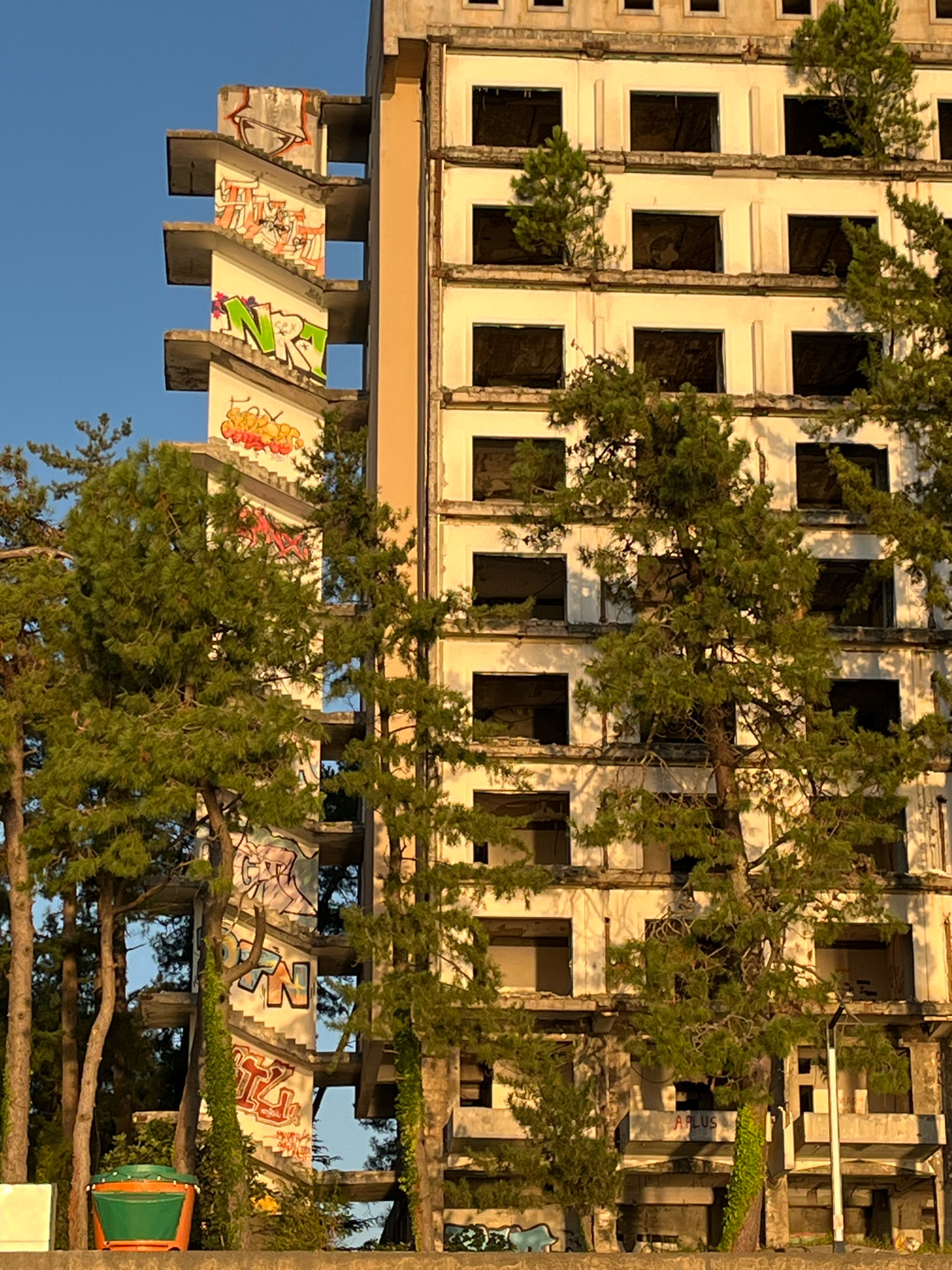 The image shows an abandoned, multi-story building with an external staircase covered in graffiti, located in Kobuleti, Georgia. The building appears to be in a state of disrepair, with many windows boarded up or empty. Trees are growing in and around the structure, obscuring parts of the facade. The sky is a clear blue, suggesting daytime. There are no people visible in the image. In the foreground, a green and orange bucket is visible. The overall impression is one of decay and overgrowth.