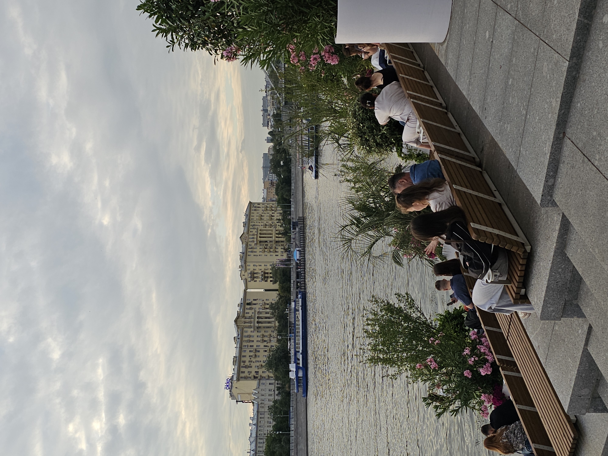 The image shows a group of people sitting on a wooden bench overlooking a wide river. The river is lined with buildings, suggesting an urban setting. The sky is overcast with pale clouds, and the light suggests it is late afternoon or early evening. The people appear to be relaxed, some looking at their phones, others conversing. The scene is peaceful and leisurely. There are trees and flowering bushes with pink flowers near the bench, adding a touch of nature to the urban landscape. A boat is visible on the river. There is text visible on one of the buildings that reads "ökko". The overall mood is tranquil and serene. The location is identified as Moscow, Russia.