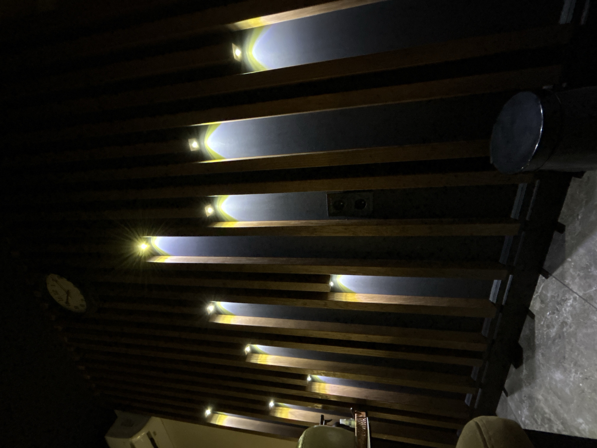 The image depicts a dimly lit interior space with a distinctive ceiling feature. The ceiling is constructed with parallel wooden slats, and recessed lights are integrated between these slats, casting light upwards. A clock is visible on the left, while a cylindrical object is on the right edge. The bottom portion of the image shows a partial view of seating furniture. The location is unknown, and there is no discernible information suggesting it is Votkinsk, Russia. There is no information about any people, subjects, actions, or interactions in this image.