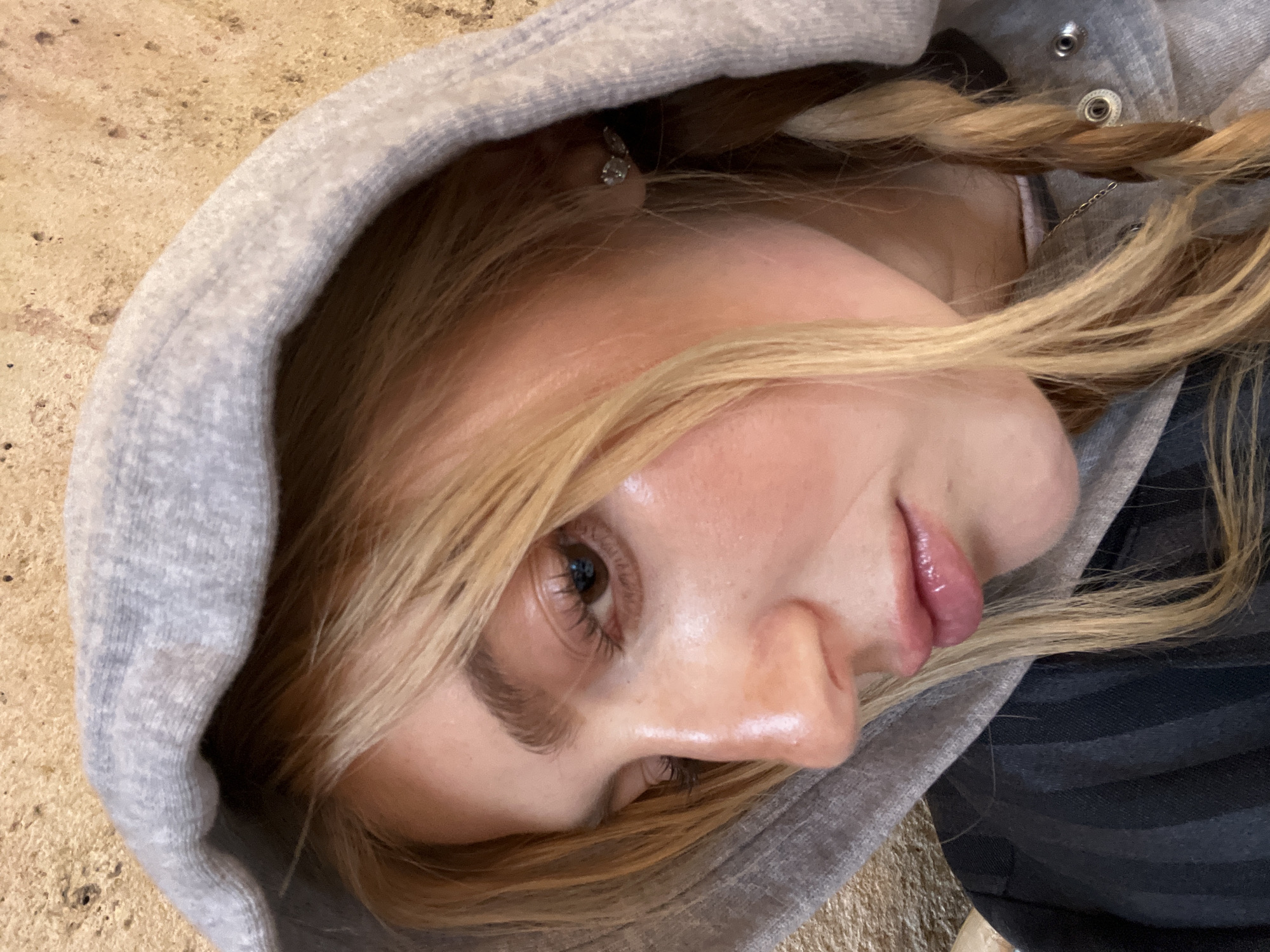A close-up, angled portrait shows a young woman wearing a gray hooded sweatshirt. Her blonde hair is styled in braids. She is wearing an earring. The background appears to be an indoor floor. The image does not provide details that would allow the location to be identified as Paris, France.