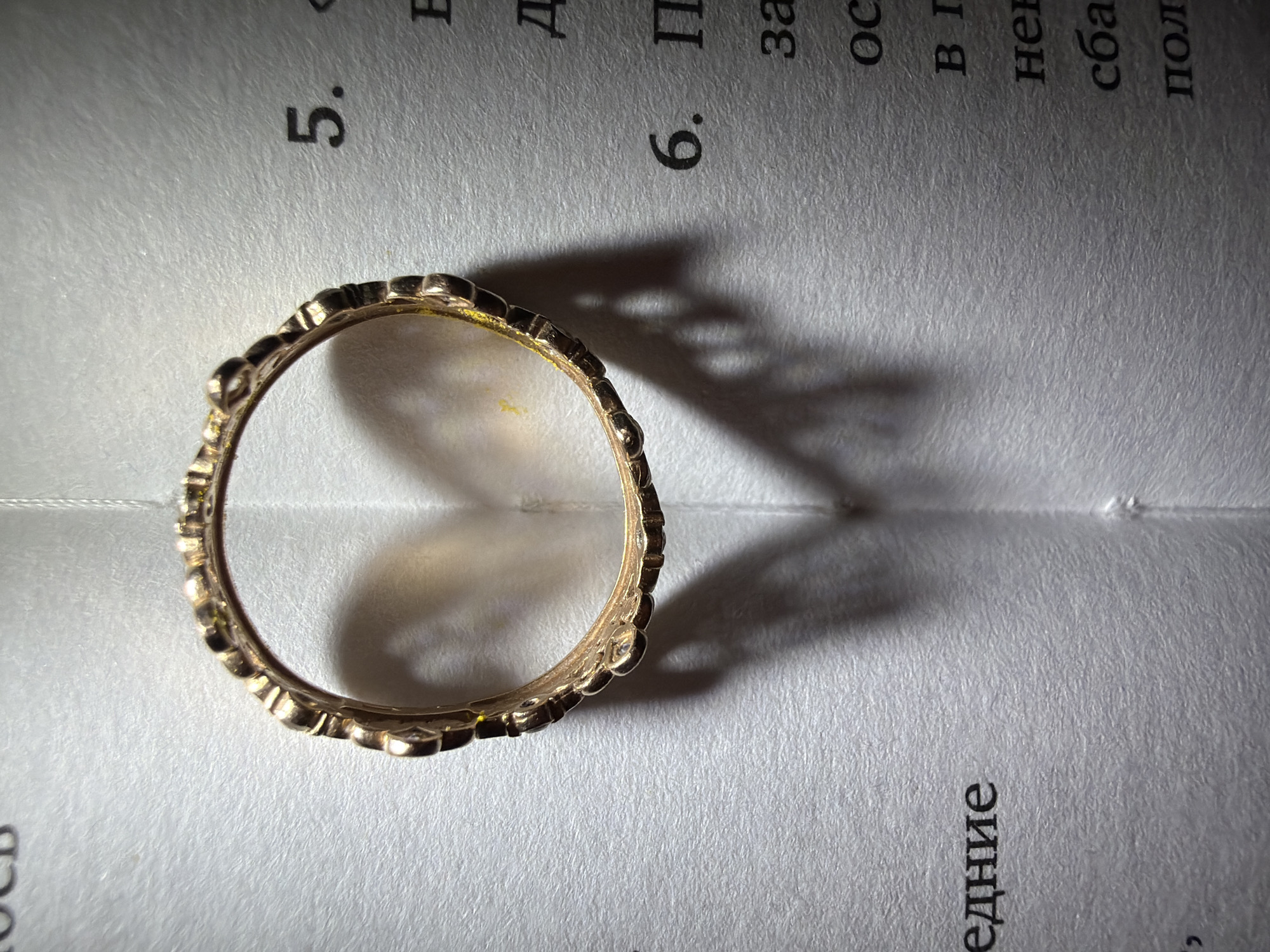 An ornate gold-colored ring is centered in the frame, resting on what appears to be two open pages of a book or thick paper. The ring has a decorative, possibly filigree or studded, design around its entire circumference. A strong, directional light source illuminates the scene from the left, casting a prominent, dark shadow of the ring onto the paper to its right. The shadow distorts slightly across the textured paper surface and reveals details of the ring's inner structure.
The paper itself is off-white or light grey with a visible fibrous texture. It has a central crease, suggesting it's an open book or folded sheets. The pages are covered in Cyrillic text. On the left page, the number "5." is clearly visible, along with fragmented Cyrillic letters above and below it. On the right page, the number "6." is prominent. Other visible Cyrillic characters and partial words include "П", "за", "ос", "в", "не", "сба", "пол" along the top and middle sections, and the word "едиие" (likely part of a larger word like "предисловие" - preface, or "сведение" - information) clearly visible at the bottom right. The text and numbers suggest a structured document, possibly a textbook or academic material. The sharp shadows indicate an indoor setting with an artificial light source. The Cyrillic script is consistent with the location context of Chaikovsky, Russia.