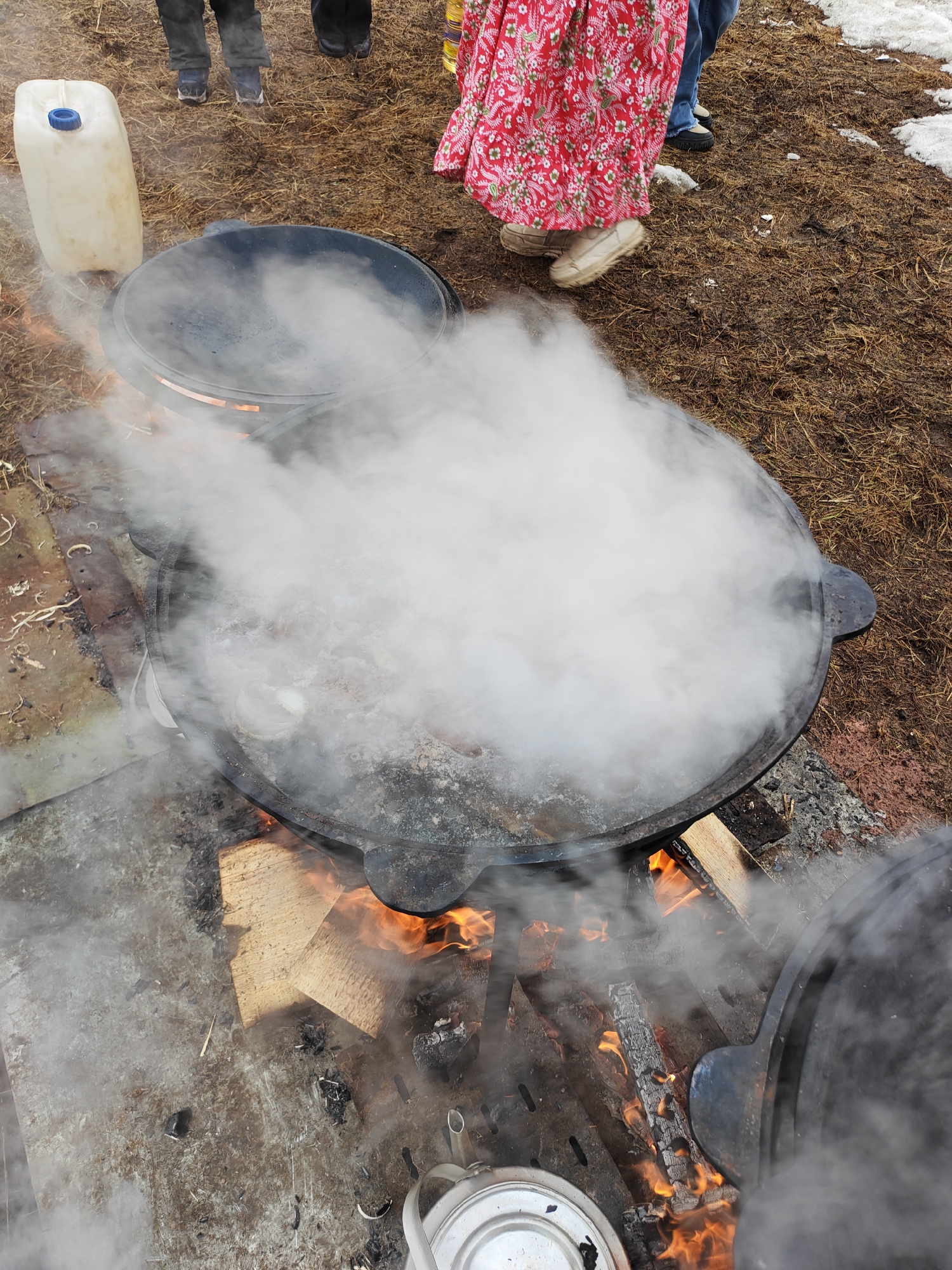 The image captures a scene outdoors in what appears to be a partially snowy environment, potentially in the city of Ugolnye Kopi, Russia.
Several people are present, as indicated by their legs and feet visible at the top of the image, with one person wearing a red floral dress.
The central focus is a large, steaming cast iron pot situated over an open fire. The fire is actively burning, fueled by wood. Another similar pot with a lid is partially visible behind the main pot, and a white plastic container is visible to the left. A white teapot is also seen in the lower-left corner.
The ground is covered with a mix of dirt, dry grass, and patches of snow. The overall impression is one of cooking or food preparation taking place in an outdoor setting.