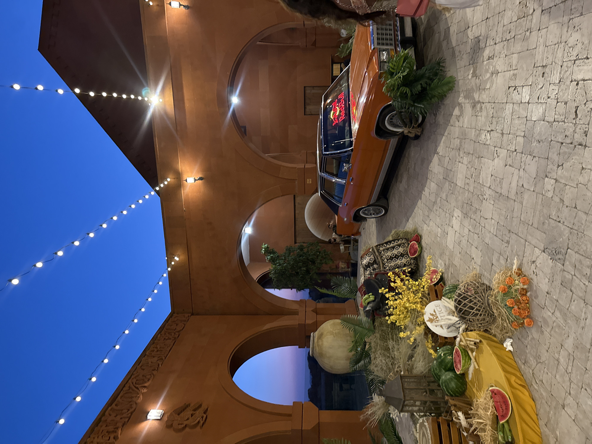 An orange vintage sedan is parked on a cobblestone surface in front of a tan stone building with arches, decorated with string lights against a twilight blue sky. The car's dashboard is illuminated by a bright red neon "Red Bull" sign. In the foreground, a display features watermelons, greenery, dried grasses, a lantern, and decorative woven spheres. To the right, the partial figure of a person in a pink garment is visible. The scene appears to be an outdoor event or gathering, potentially in Sasunik, Armenia, given the context.