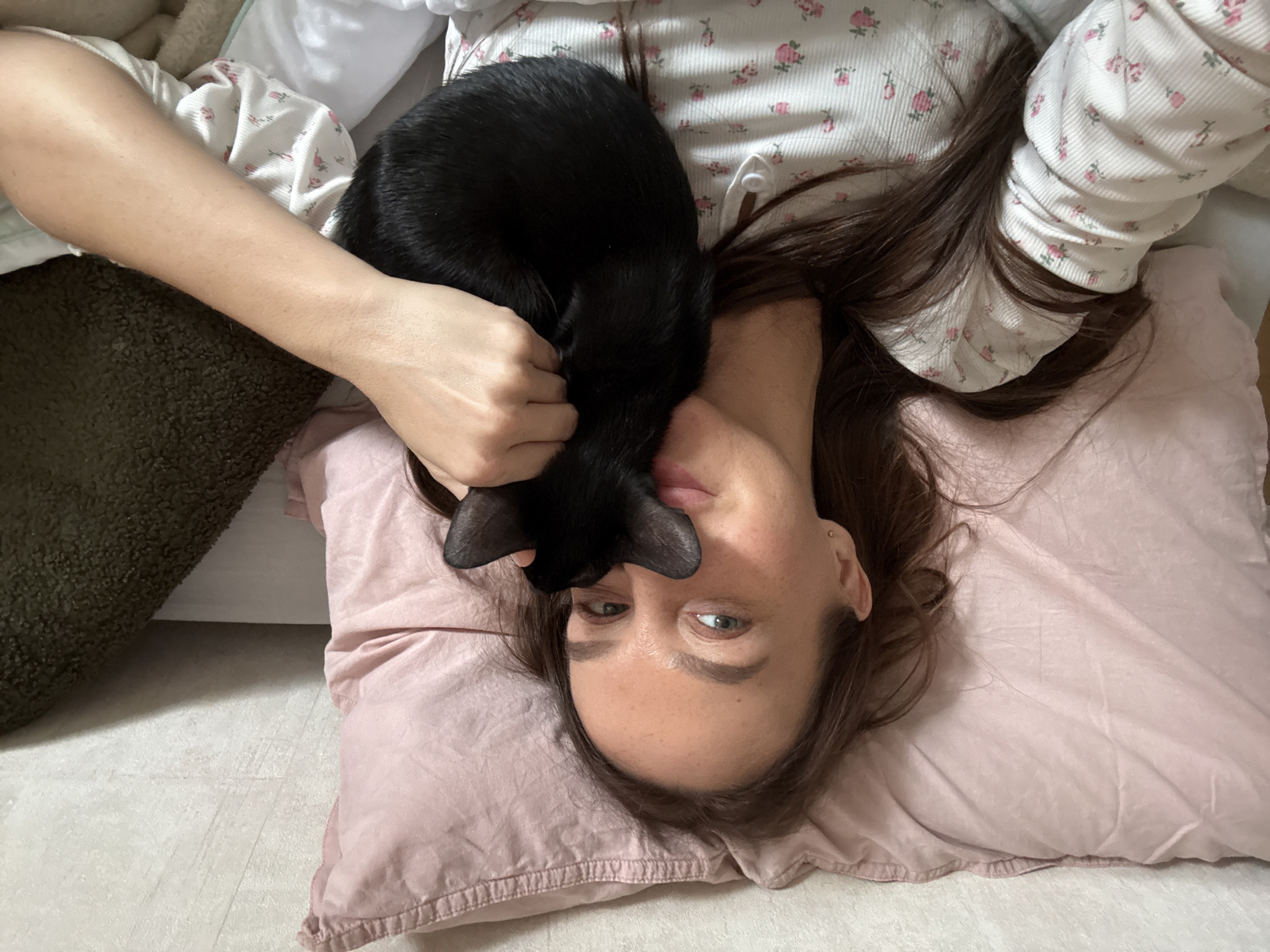 An overhead view shows a young woman lying on her back, her head resting on a light pink pillow. Her long, dark hair is fanned out around her head and shoulders. She is wearing a white, ribbed long-sleeved shirt with a repeating pattern of small pink roses. A black cat is curled up on her chest, its head nestled close to her mouth and nose. The woman's right hand is gently placed on the cat's back. She looks directly at the camera with a neutral, slightly content expression, her light-colored eyes open. The cat appears relaxed or sleeping. The background consists of light-colored flooring or bedding, with a dark green textured cushion or blanket visible on the left side of the frame. This intimate scene depicts a quiet moment of affection and comfort between the woman and her feline companion, likely taking place indoors in a cozy setting in Chaikovsky, Russia. There is no visible text in the image.