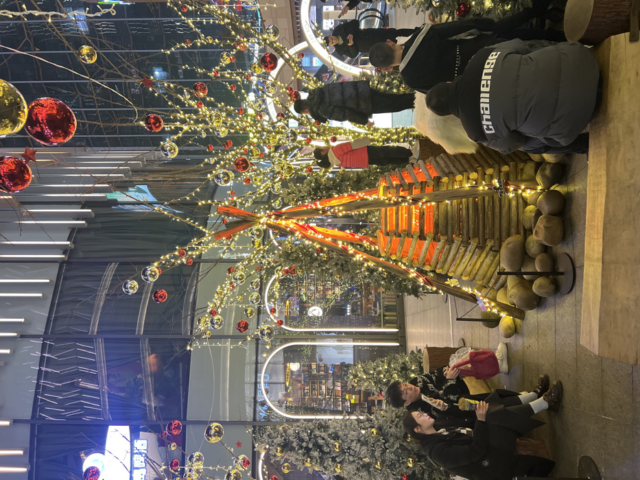 The scene depicts an illuminated indoor public space within a commercial building in Liangjiang, China, decorated for a festive occasion. Centrally located is a large, conical structure composed of stacked light-colored logs, resembling a teepee, internally illuminated with orange and yellow lights to simulate a fire. This structure is surrounded by multiple artificial evergreen trees, some flocked with a white material, and numerous bare branches, all extensively adorned with warm white string lights and red and gold spherical ornaments. Large red and gold reflective spherical ornaments are suspended from the ceiling.
In the foreground left, a man and a woman are seated on a low wooden platform. The woman, in a dark jacket, holds a beverage cup and looks at a mobile phone. The man, wearing a dark patterned top and glasses, also interacts with a mobile phone. A red tote bag rests on the platform between them.
In the midground, several individuals are present. A child wearing a pink jacket stands on a circular wooden stump amidst the decorated trees, while an adult male in a dark puffer jacket stands nearby. Further to the right, a man wearing a grey and black puffer jacket with "Challenge" written in white across the back is crouched down, facing away from the camera. Adjacent to him, another man in a black top with a white strap over his shoulder stands.
The background features large glass walls or windows reflecting interior lights and vertical strip lighting on the building's structure. Two prominent arched doorways, outlined with bright white lights, lead into interior retail spaces that appear to be a bookstore or shop with visible shelves. An escalator is discernable in the far right background. The environment is brightly lit by artificial sources.