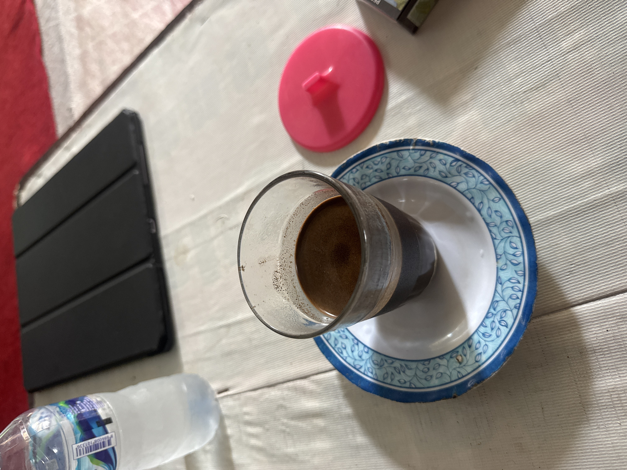 The image depicts a close-up view of a table setting. In the foreground, a glass of dark liquid, presumably coffee, sits on a small plate with a blue and white floral pattern. To the right of the coffee cup, a bright pink lid with a central knob is placed on the table. Further to the right, a portion of a cigarette pack is visible. In the background, a black tablet device lies on its side. To the left of the coffee cup, a partially filled bottle of water with a blue and white label is present. The table is covered with a light-colored textured cloth, and a red surface is partially visible in the upper right corner. The scene is lit by natural light, casting shadows on the table. The overall impression is a casual, indoor setting, likely a home or a cafe, in Busungbiu, Indonesia.