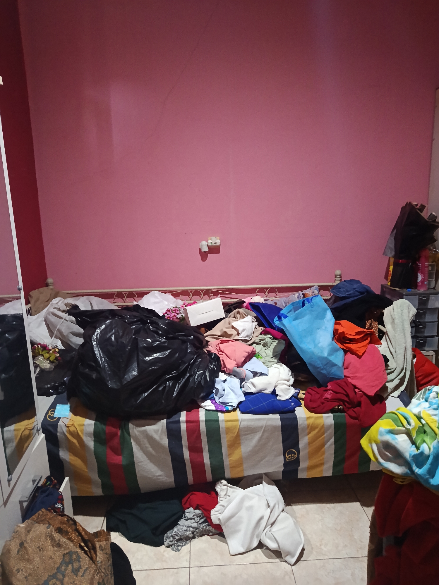 The image depicts a cluttered bedroom with a pink wall. A bed, covered with a colorful striped comforter, is laden with a large black trash bag filled with items, along with numerous piles of clothing in various colors and states of disarray. To the right of the bed, plastic storage drawers are stacked, holding more assorted items. On the floor in front of the bed, more clothes are scattered. A white cabinet with a mirrored door is visible on the left side of the frame. The scene is dimly lit, suggesting it is nighttime or the room is not well-lit. The overall impression is one of disorganization and clutter. The specific location in Keboananom, Indonesia, is not visually represented in the image.