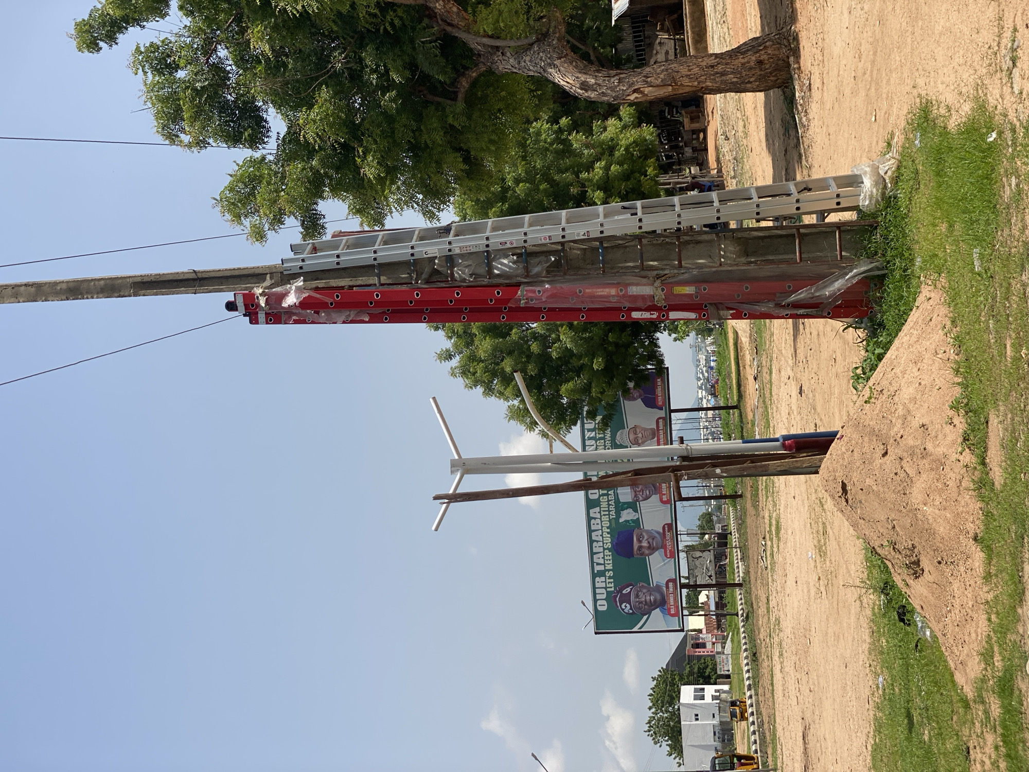 This media file captures an outdoor scene in Jalingo, Nigeria, on what appears to be a clear and sunny day. The primary focus is on several ladders, both silver and red, stacked and attached to a pole, likely for transport or storage. Behind the ladders, a large tree with lush green foliage provides a natural backdrop.
In the mid-ground, a series of large billboards with political messaging is visible. These billboards feature portraits of individuals, suggesting a campaign or electoral event. Text on the billboards includes phrases like "OUR TARABA O LET'S KEEP SUPPORTING T TARABA" and names such as "BOLA AHMED TINUBU", "DR. AGBV", and "ILIYA KEFAS ALN". The presence of these political advertisements strongly indicates the location is in Nigeria, specifically in the Taraba region.
The ground is covered in sandy soil, with patches of green grass on the right side of the frame. A large, rough, earthen mound is also present in the foreground. In the distance, some buildings and vehicles can be observed, hinting at a semi-urban or roadside setting. The overall impression is one of a vibrant, possibly bustling, outdoor environment, with a clear emphasis on the political landscape through the prominently displayed billboards.