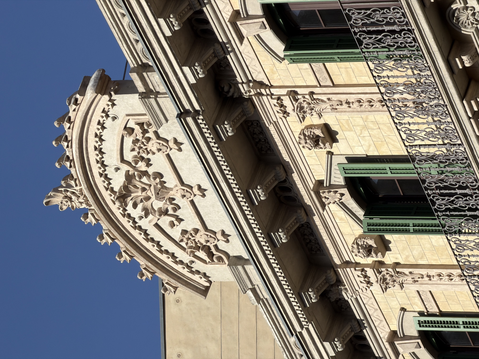 The image shows a close-up view of the upper facade of a building in Barcelona, Spain, under a clear blue sky. The architecture features ornate stone detailing.
The top of the building is crowned with a decorative pediment, intricately carved with floral motifs and a central crest. Below this, a prominent cornice runs horizontally, supported by ornate corbels that are themselves decorated with carved faces resembling lions. Further down, there is a balcony with a wrought-iron railing featuring elaborate scrolling patterns. This balcony overlooks windows with green shutters. The facade is constructed from light-colored stone blocks, likely sandstone or a similar material, giving it a warm, sun-drenched appearance. The building's style suggests a historical period, possibly Art Nouveau or a similar ornate European architectural movement. The lighting indicates a sunny day, with strong shadows highlighting the sculpted details.
