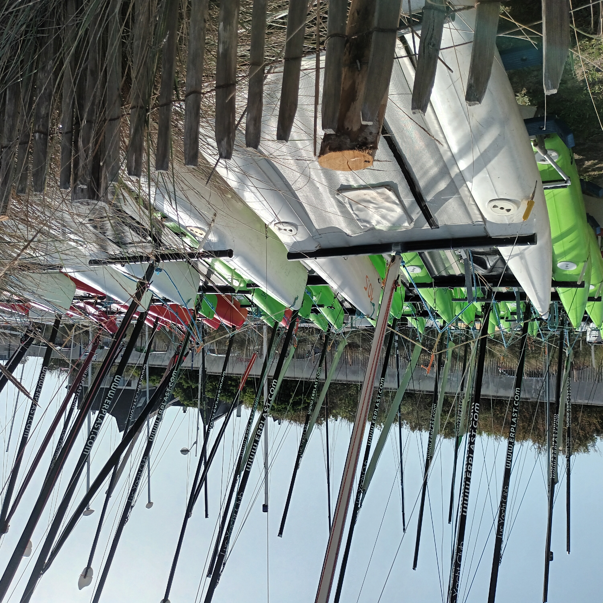 The image shows a row of small sailboats, likely catamarans, stored on a rack. The hulls of the boats are primarily white and green, with some red boats also visible. The masts are black and have the words "www.ERPLAST.COM" and "Luc Yacht Club" printed on them. The boats are stored in a beach club with a wooden fence made of untreated wood. The area is likely near the water, as the image features light-colored skies. The location is Unknown, Unknown.