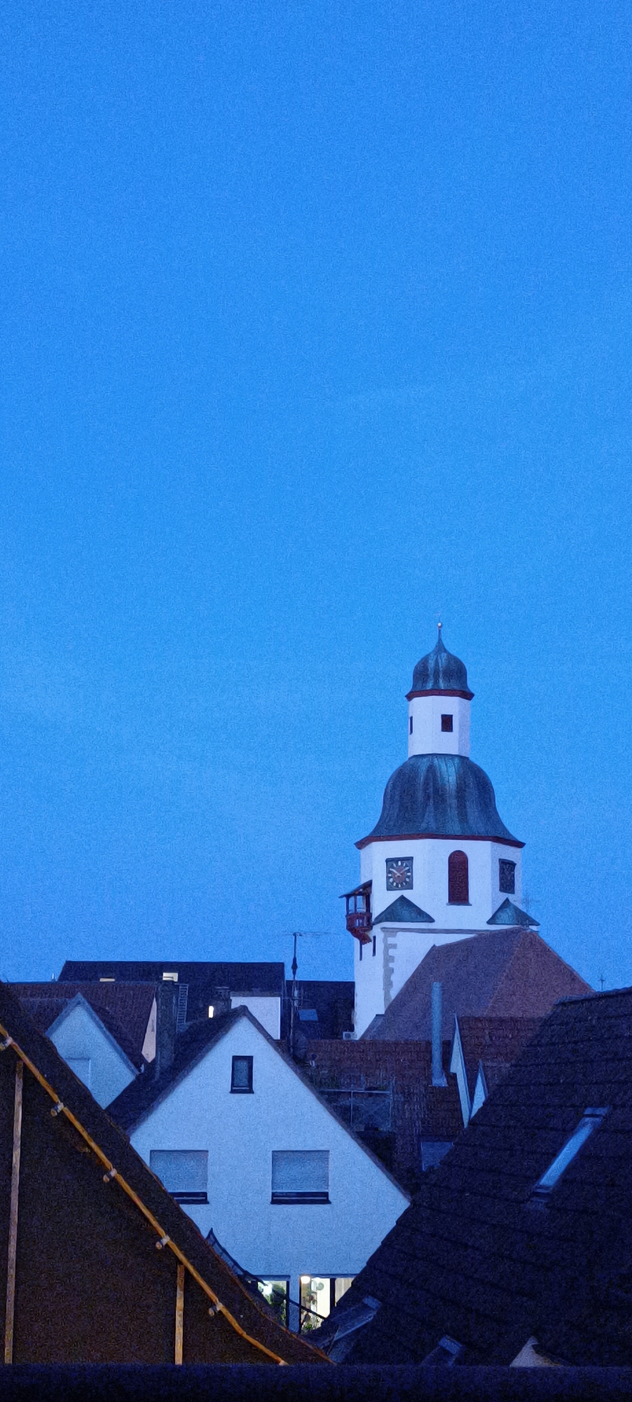 The image shows a low-angle, vertical shot of a town in Winnenden, Germany, taken at twilight or dawn. The sky is a deep, cloudless blue. In the center right of the frame, a white church tower with a dark, domed roof rises above a cluster of houses. The tower has a clock face with Roman numerals and a small red balcony. The roofs of the surrounding houses are dark and tiled. The overall scene is peaceful and quiet, suggesting a residential area or town center.