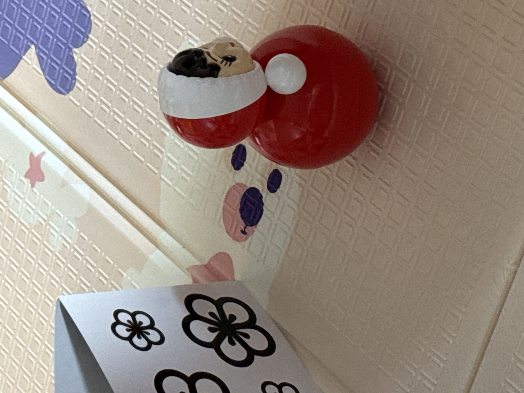 The image depicts a red Daruma doll with a white eye and an attached figurine head wearing a white headband. The Daruma doll is positioned on a white patterned surface, likely a wall, with a cartoon bear face and a paper card with floral designs underneath. The environment seems to be indoor, possibly a room with patterned walls and decorative elements. The specific location is not identifiable based solely on the image.