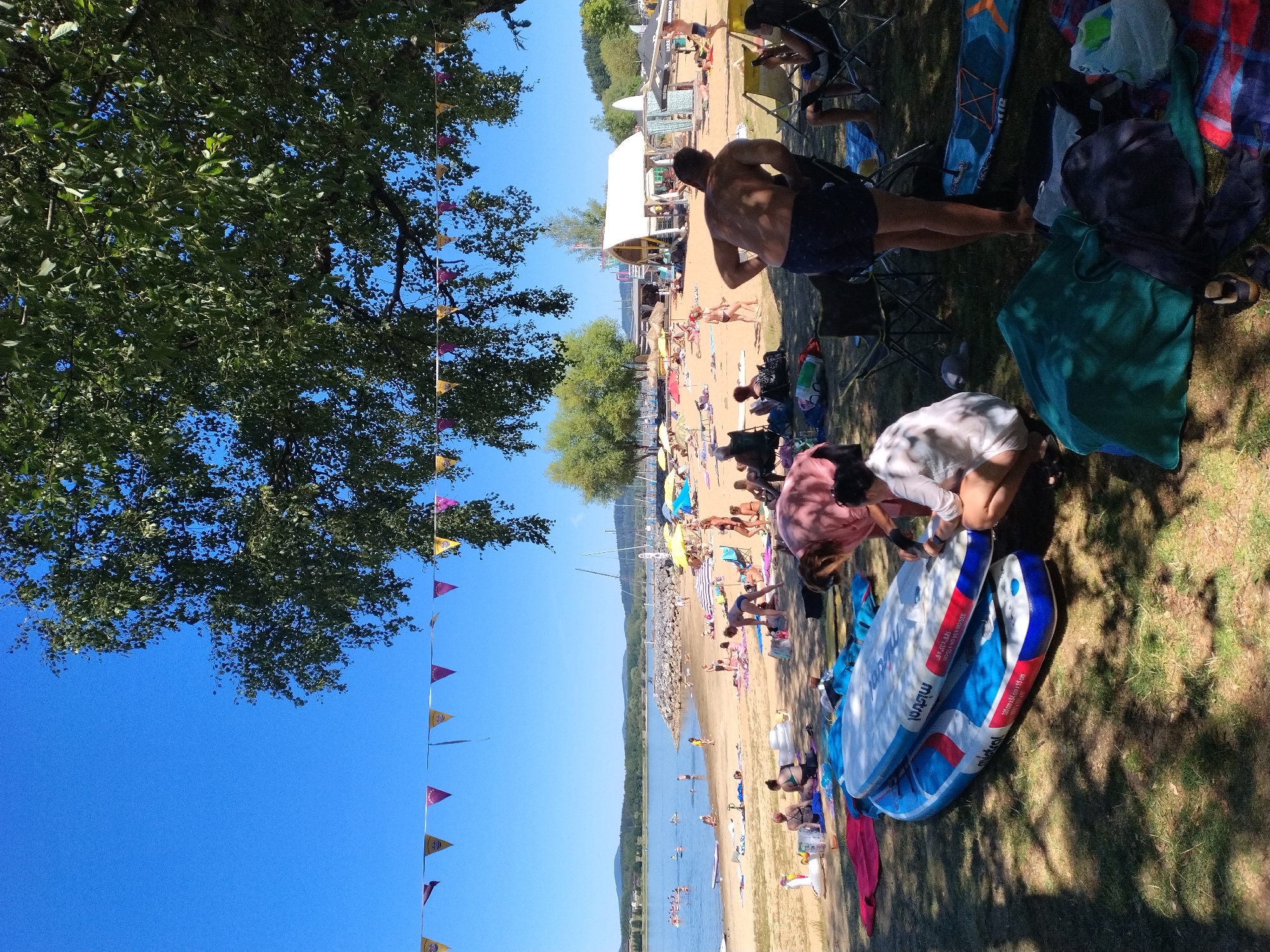 This is a wide shot of a crowded beach on a sunny day, with a lake and hills in the background. The foreground shows several people relaxing on the grass under the shade of a large tree, with several stand-up paddleboards and beach gear laid out. A string of triangular flags is visible in the upper portion of the image. In the middle ground, people are spread out on the sandy beach, some sunbathing, others swimming in the lake, and some are near a beach bar. The scene appears to be a popular recreational area, likely a lake beach in Radslav, Czechia, judging by the typical amenities and natural setting.