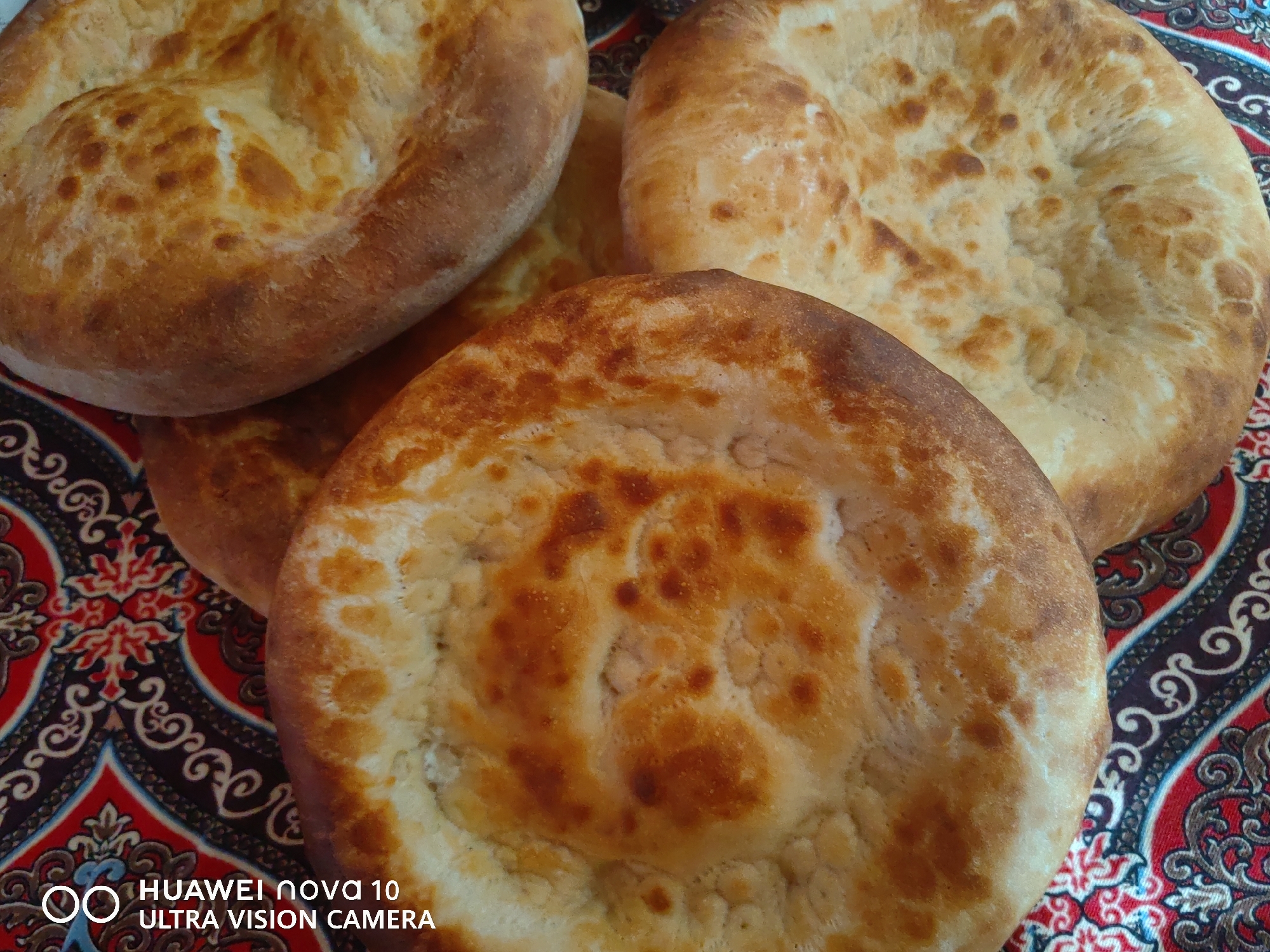 In Jetim-Dobo, Kyrgyzstan, four pieces of freshly baked flatbread, likely lavash or non, are arranged on a red and black patterned textile. The bread is golden brown with darker browned spots and has a slightly puffed, uneven surface. The textile features an intricate paisley or floral design. The bottom left corner of the image displays a Huawei Nova 10 Ultra Vision Camera watermark.
