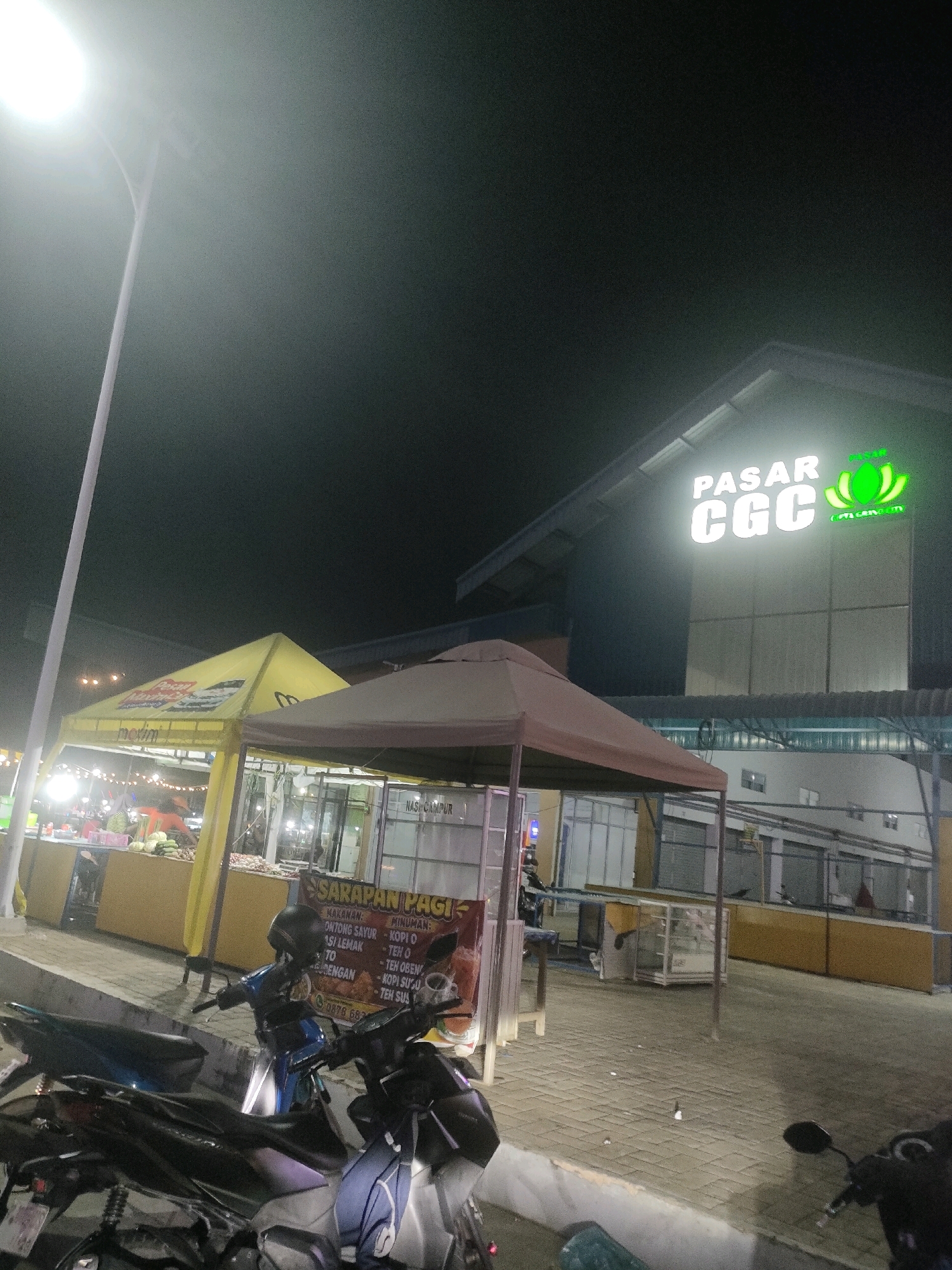 An outdoor night scene depicts a food market or outdoor dining area in Batam, Indonesia. In the background, a building with a large illuminated sign reading "PASAR CGC" is visible. In the mid-ground, two market stalls are set up with yellow and brown awnings, illuminated by string lights and overhead lighting. One stall has produce displayed, including what appear to be melons. A sign on the stall reads "SARAPAN PAGI" (Breakfast) with listings for food and drinks. In the foreground, several motorcycles are parked on a paved walkway. The overall atmosphere is bustling and commercial, with evidence of food service and retail activity. The lighting suggests it is nighttime.