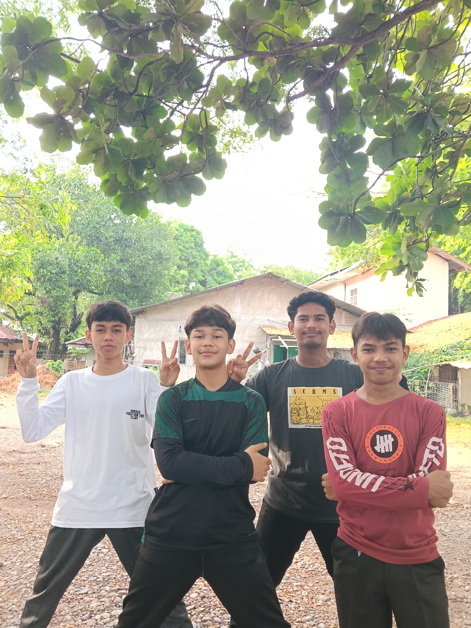 Four young men are standing outdoors, under a tree with broad leaves. They are posed together in a gravelly yard in front of several houses of varying construction styles. The first young man from the left is wearing a long-sleeved white shirt and grey pants, holding up the peace sign with his left hand. The second young man is wearing a long-sleeved dark green and black shirt, with his arms crossed. The third young man is wearing a short-sleeved grey t-shirt. The fourth young man is wearing a long-sleeved reddish-brown shirt with writing down the sleeves, and has his arms crossed. The houses in the background are a mix of concrete and wooden structures. The picture was taken outdoors during daylight hours. The location is Lhokseumawe, Indonesia.