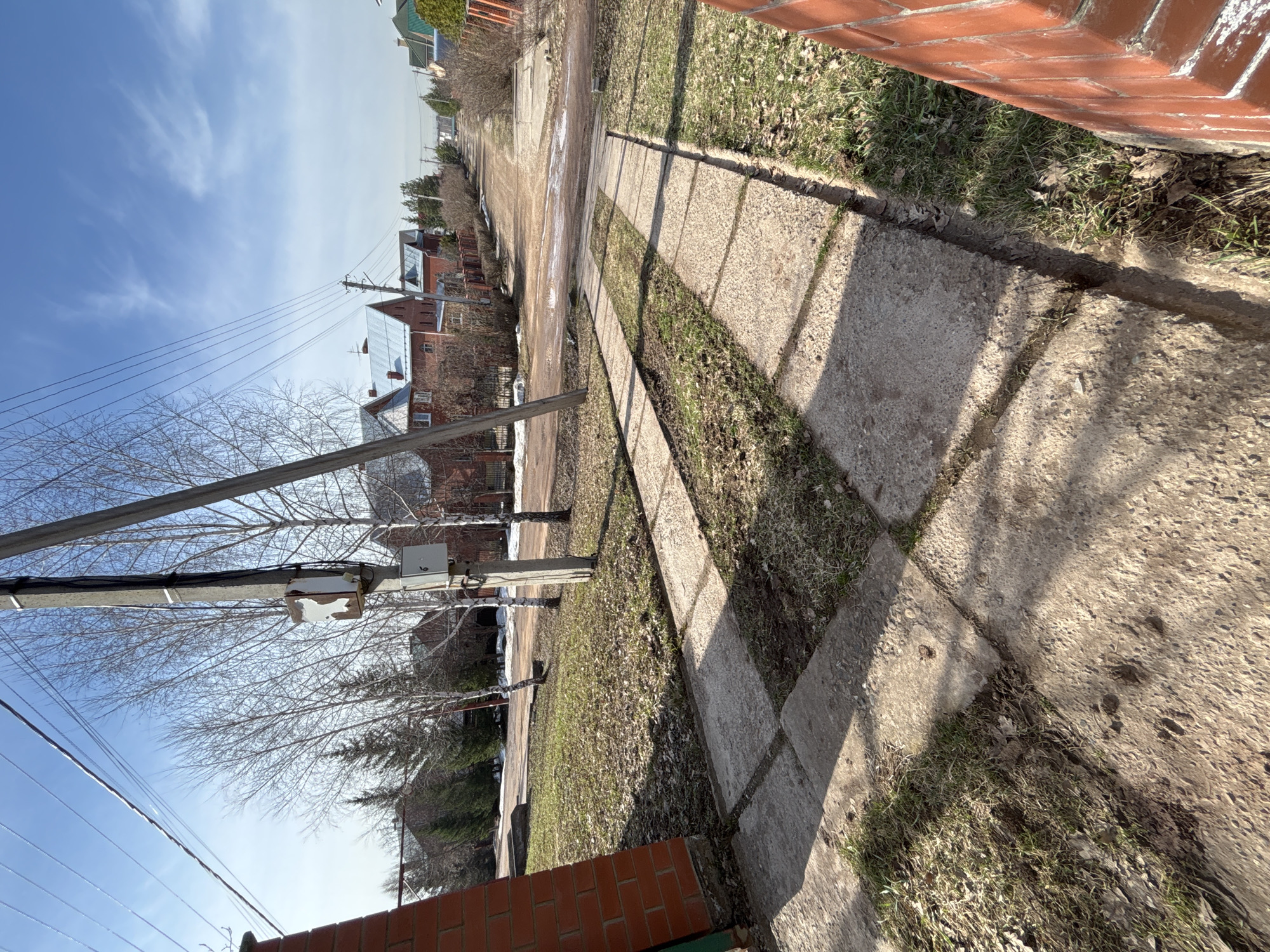 The image shows a street scene in Ферма, Russia. The foreground features a concrete sidewalk bordered by a brick wall on the right and grass on both sides. A utility pole with wires runs along the left side of the street. The street itself appears to be a dirt road, stretching into the distance. Houses are visible along the street, suggesting a residential area. The sky is blue with scattered clouds. The image captures a common street scene in the area, emphasizing the mix of paved and unpaved surfaces, the presence of utility infrastructure, and typical housing.