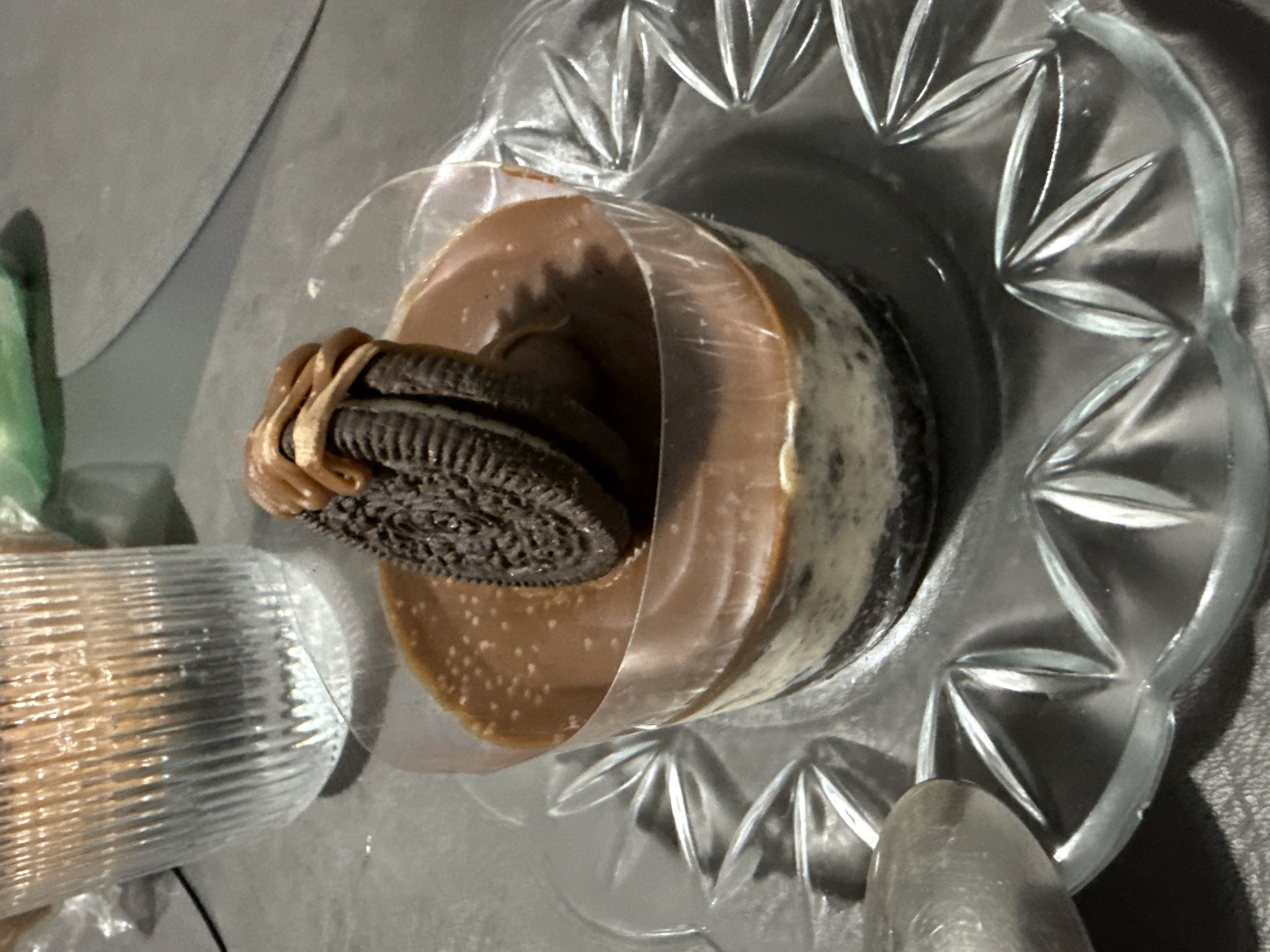 A dessert sits in a clear, decorative glass dish. The dessert appears to be composed of layered elements, including a brown, glazed topping, a lighter-colored base, and an Oreo cookie positioned on top. The dish is partially encased in a clear plastic wrapping. A ridged glass object and a spoon are partially visible to the side of the dish. The surface underneath is a gray, textured material. There is no indication of location. There are no people visible.