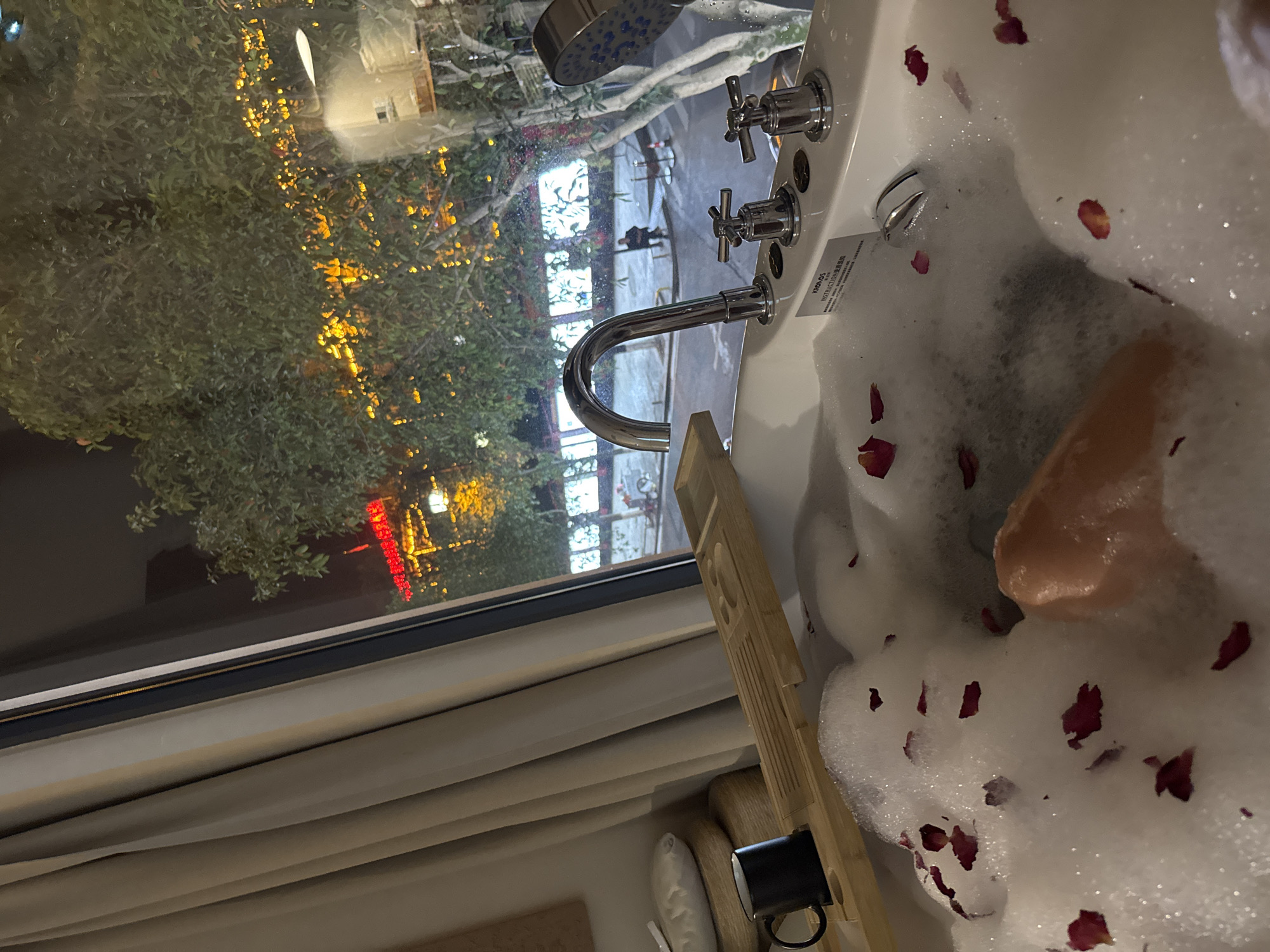 The image depicts an indoor scene at night, overlooking a city street in Wulingyuan, China. The primary subject is a white bathtub filled with dense white foam bubbles and scattered red rose petals, with a pale limb, presumably a human leg, visible submerged within the bubbles. A chrome gooseneck faucet, two chrome cross-handle control knobs, and a chrome handheld showerhead with blue-toned nozzles are mounted on the tub's rim. A label bearing "KRONOS" and "INSTRUCTION" in both English and Chinese characters is affixed to the tub surface.
To the immediate left of the tub, a light-colored wooden bath caddy spans the width, holding a black ceramic mug. Further left, a white pillow rests on stacked woven wooden stools, positioned next to partially drawn light-colored curtains.
Beyond a large window, a nighttime urban environment is visible. The street appears wet, reflecting overhead light. Trees with green foliage are illuminated by numerous small, warm yellow lights. A red neon sign with Chinese characters is visible on a building across the street. On the sidewalk, a solitary individual is seated, while another person walks further down the street. A red and white striped barrier or cone is also present on the sidewalk. The ambient lighting from the street and buildings casts a warm glow, with some interior reflections visible on the windowpane.