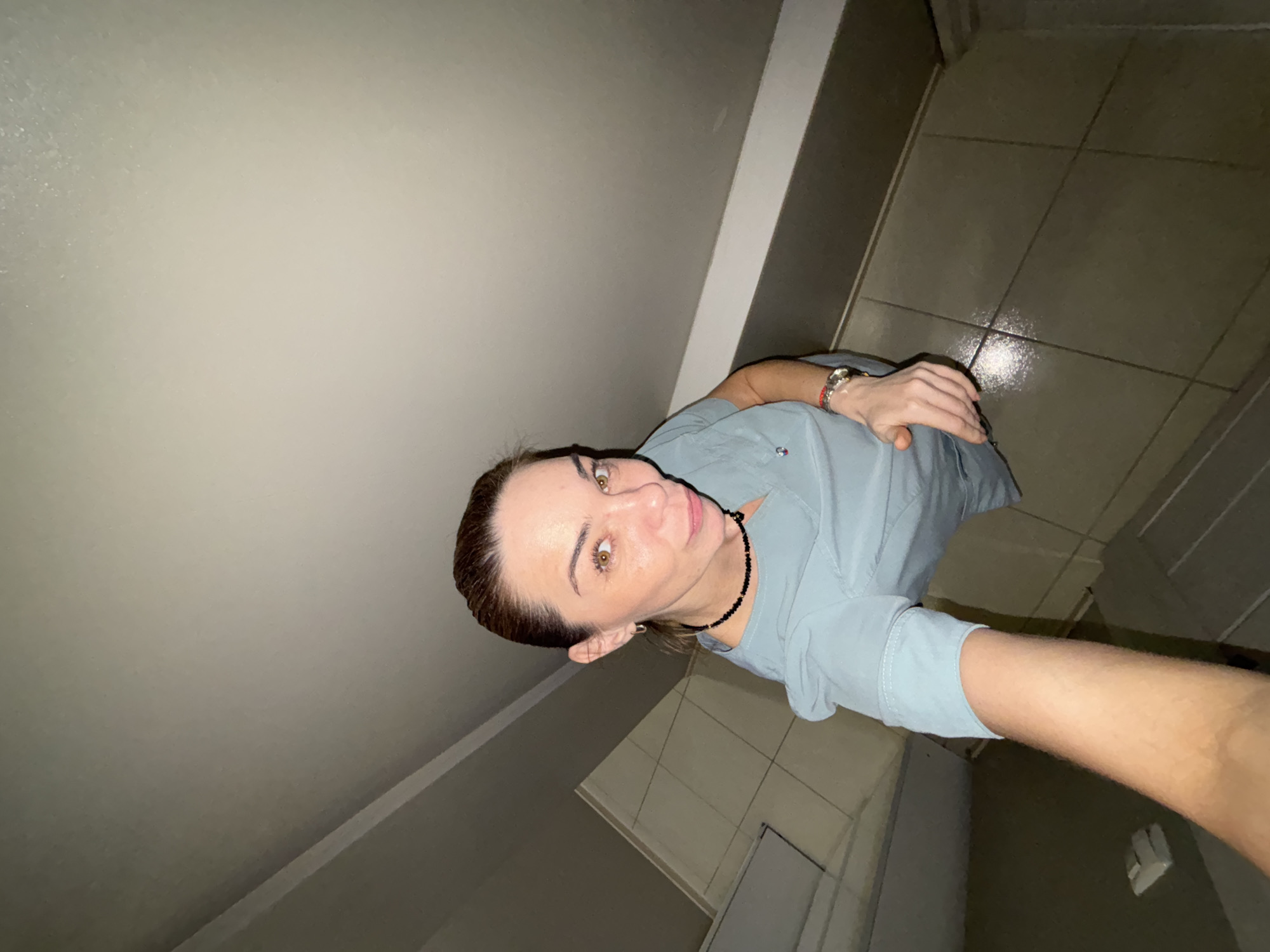 A woman is taking a selfie in a tiled hallway with a low ceiling. She is wearing a light blue scrub top, a black beaded necklace, and a watch on her left wrist. Her hair is pulled back, and she is looking at the camera with a slight smile. The hallway appears to be in a building, possibly a hospital or clinic, given the uniform. The floor is tiled and appears to be wet in some areas, reflecting the overhead light. The walls are also tiled in a lighter color. The overall lighting is dim. The location is specified as Sinyavino, Russia, but this information is not visually verifiable from the image itself.
