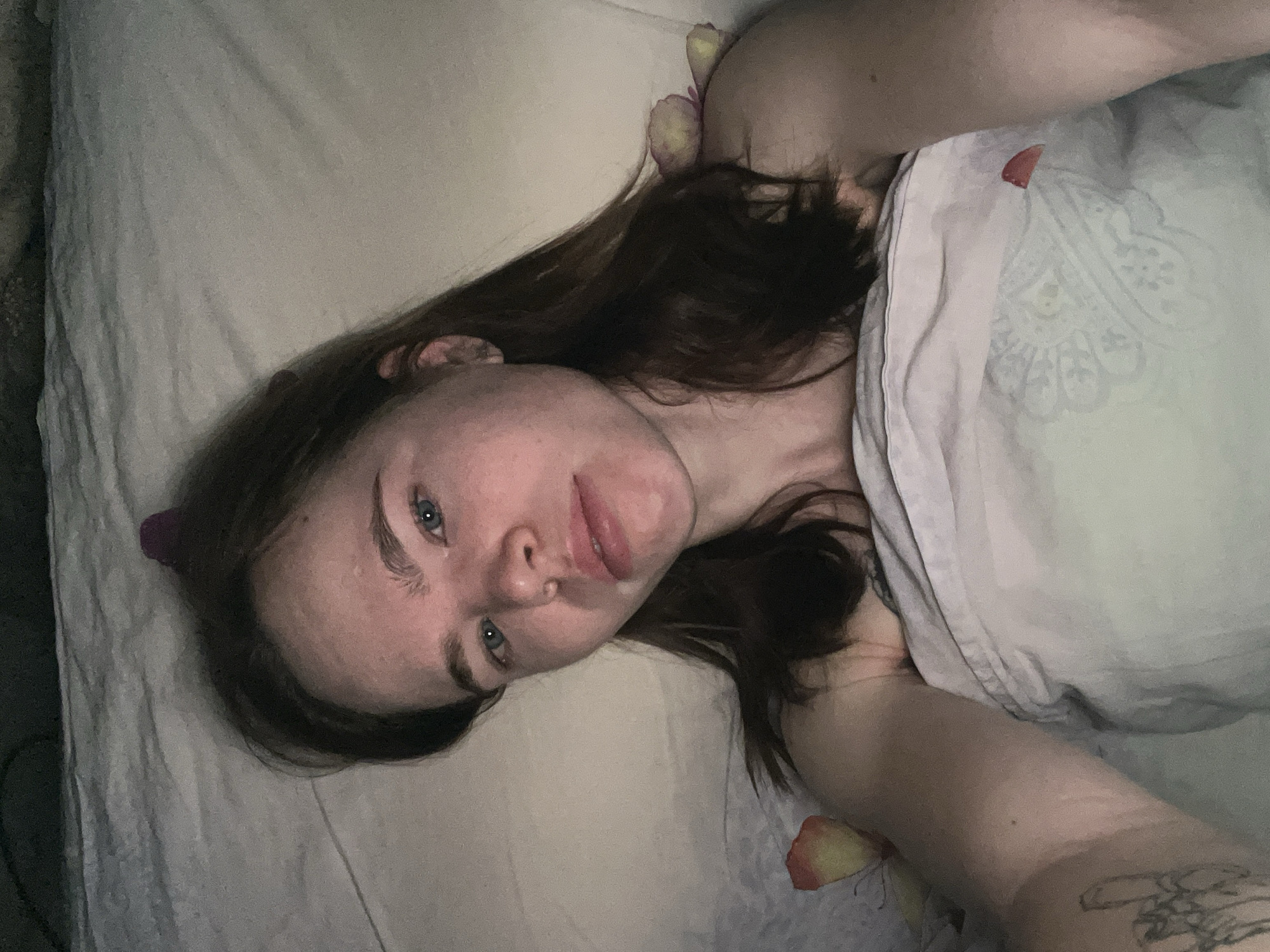 A young woman is lying on a bed covered in light-colored sheets and bedding. She has long brown hair and blue eyes, and her face is visible. Rose petals are scattered around her head and shoulder area on the bedding. Part of a tattoo is visible on her right forearm. The location, Chaikovsky, Russia, cannot be confirmed from the image.