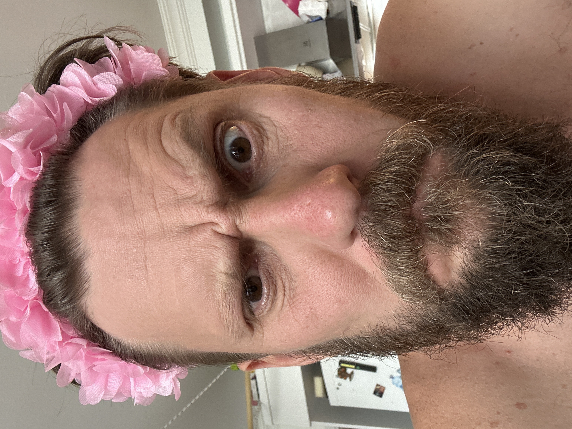 A close-up, slightly angled shot features a Caucasian male with a substantial brown and grey beard, wearing a pink floral headband. His expression appears furrowed, with one eye squinted and wrinkles on his forehead. Behind him, a section of a white refrigerator is visible, along with a light-colored wall and partial view of a doorway. The background is generally nondescript and offers no location cues, and therefore the location in the city of Fethiye, Turkey cannot be determined from the image.