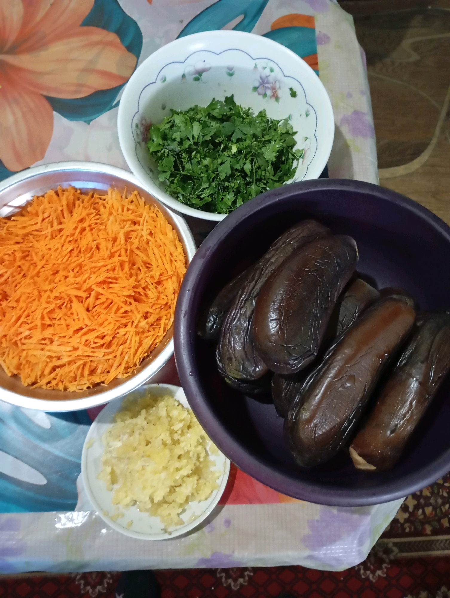 In Horishni Plavni, Ukraine, a still life photograph captures various ingredients for a meal arranged on a table covered with a patterned cloth. In the upper left, a white bowl contains finely chopped green herbs, possibly parsley or cilantro. Adjacent to it, a shallow metal bowl is filled with shredded orange carrots. To the right of the carrots, a dark purple bowl holds several cooked eggplants, their skins wrinkled and dark. In the lower right quadrant, a small white plate displays finely minced garlic. The background features a colorful floral pattern on the tablecloth. The lighting suggests an indoor setting, likely a kitchen or dining area.