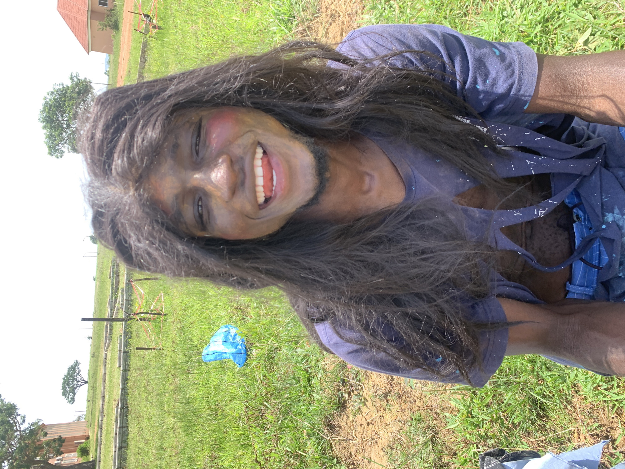 The image shows a close-up of a person, likely male, with dark, long, messy hair. Their face is covered in black paint, with streaks of red on the cheeks, resembling makeup. The person is smiling broadly with their mouth open, showing teeth and tongue. They are wearing a dark blue t-shirt that is splattered with white paint. The background appears to be an outdoor, grassy area with some construction or development in progress, indicated by wooden stakes and tape. In the distance, there are buildings with red roofs, one of which resembles a house. The overall setting suggests a potentially festive or artistic event, possibly a cultural celebration or a performance, taking place in an open, rural or semi-rural location within Gwafan, Nigeria. The bright sunlight suggests it is daytime.