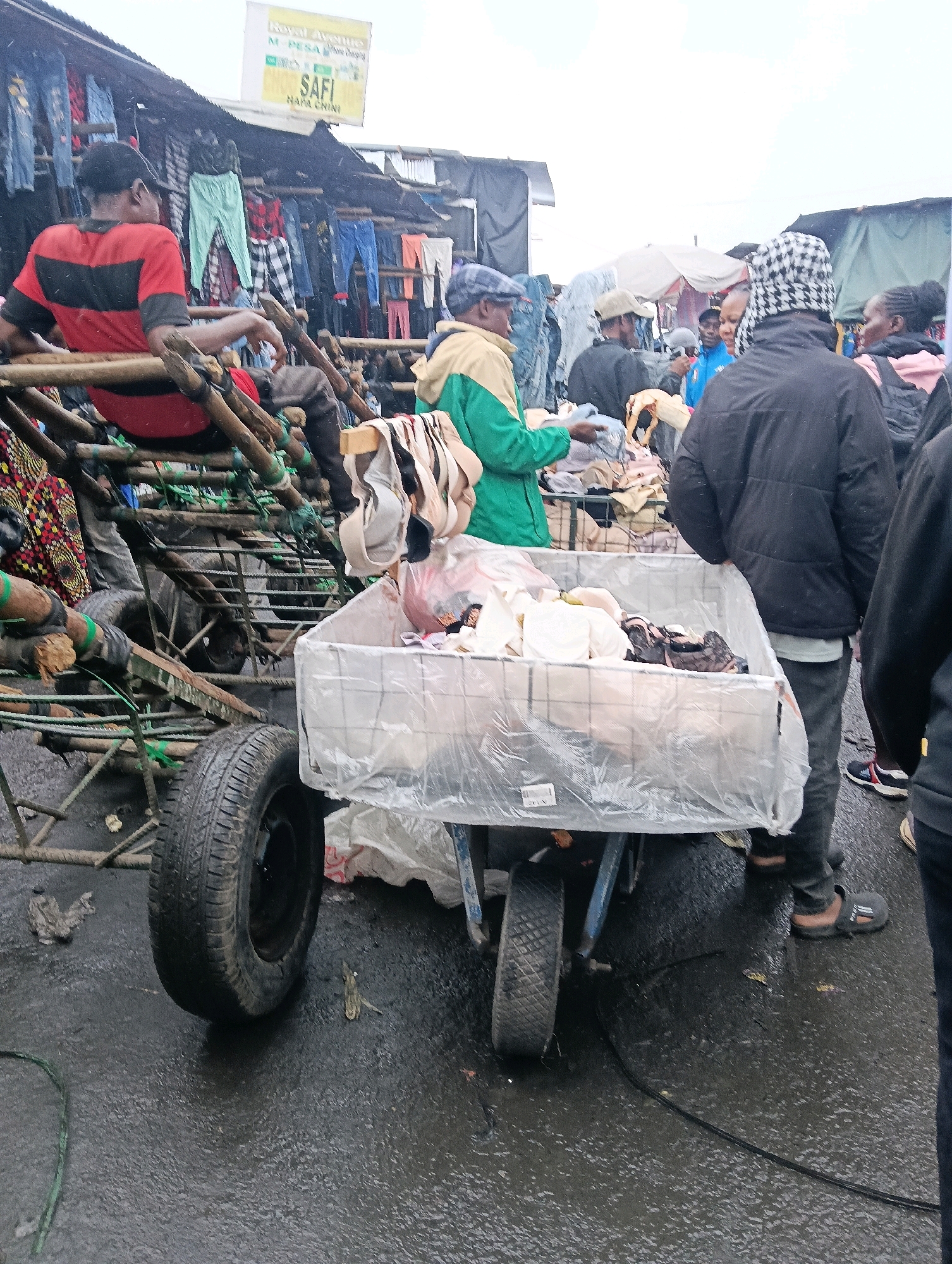 The image depicts an outdoor market scene, likely in Nairobi, Kenya, given the presence of a Safaricom M-Pesa sign. The ground is wet and appears to be asphalt, suggesting recent rain. In the foreground, a trolley is laden with a variety of clothing items, including what appear to be bras and underwear, all covered in clear plastic sheeting. A man wearing a black checkered hat and a black jacket stands behind the trolley, facing away from the camera. To the left of the trolley, a man in a red and black striped shirt sits on a wooden structure, also facing away. The background shows a market stall with clothes hanging from a rack. Several other people are visible in the mid-ground and background, some appearing to be customers or vendors. The overall impression is of a busy, possibly humid, outdoor marketplace.
