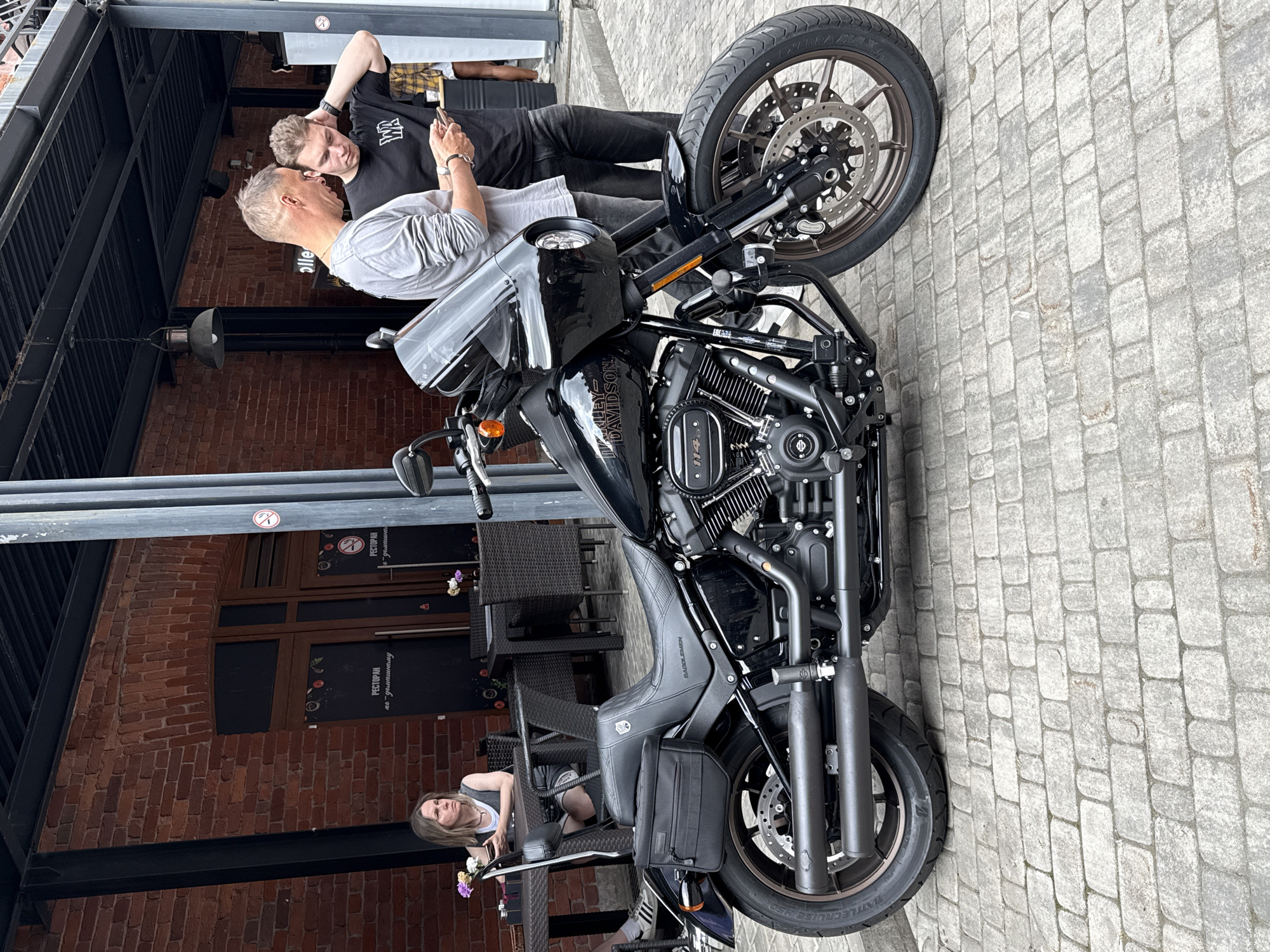 A black Harley-Davidson motorcycle is parked on a cobblestone street in front of a brick building. Two men are standing next to the motorcycle, looking at a phone. A woman is sitting at a table to the left, also looking at a phone. The building appears to be a restaurant with outdoor seating. The scene is likely Saint Petersburg, Russia, given the presence of Russian text on the building.