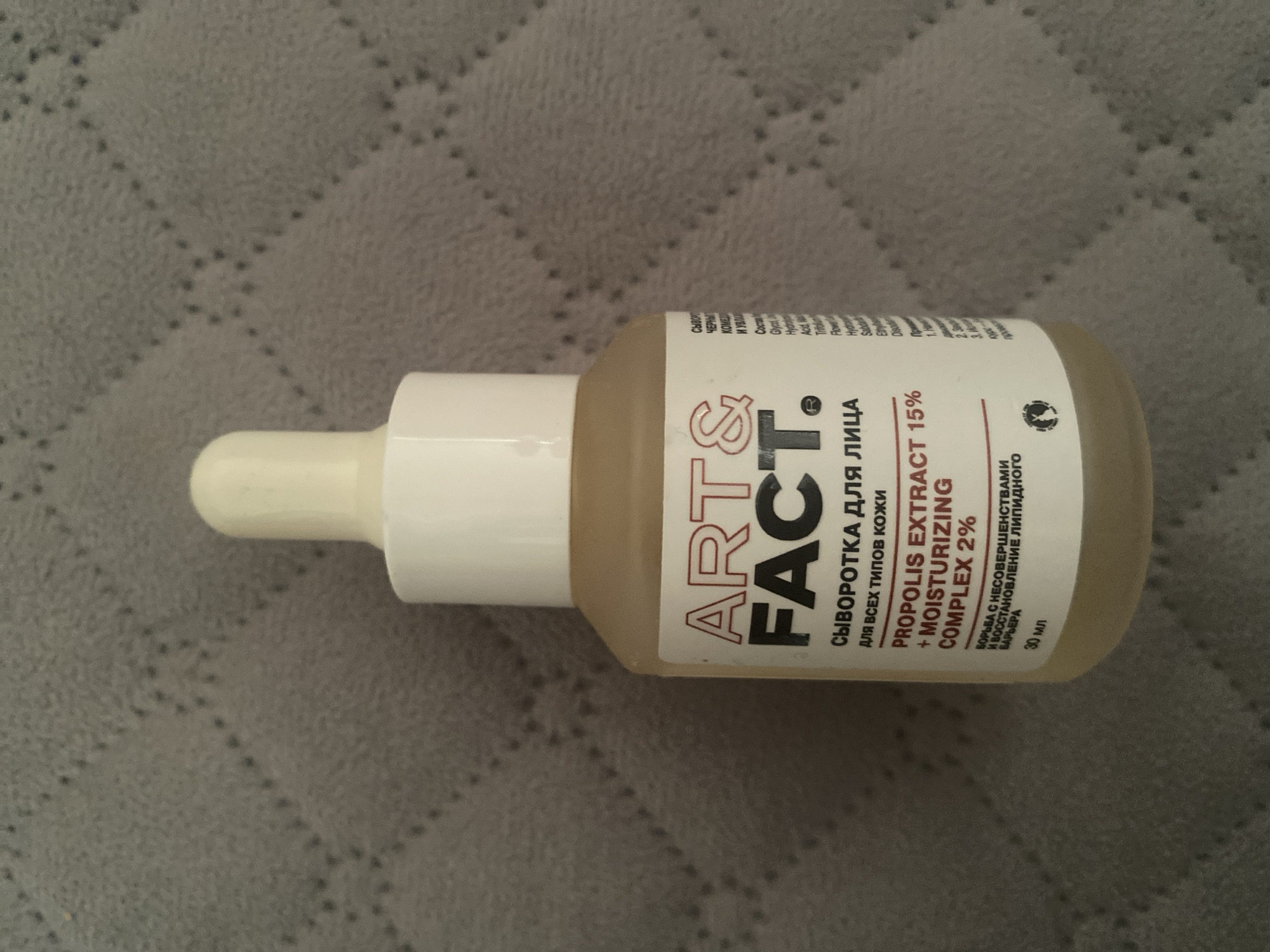 The image shows a bottle of ART&FACT serum with propolis extract and a moisturizing complex. The bottle is light brown with a white cap and dropper. The label is white with black and red text. The bottle is sitting on a textured gray surface. The location in Kizilyurt, Russia is not provided or evident from the image.