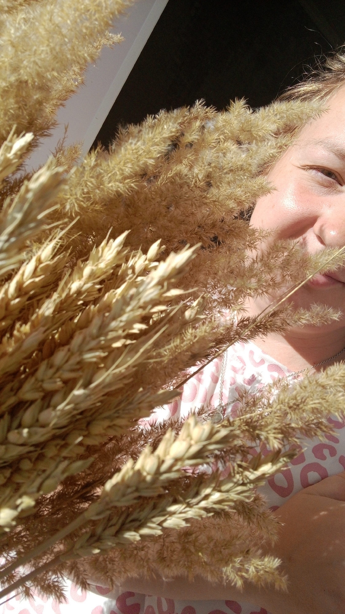 A person holds a bouquet of dried wheat and grass in front of their face. The scene appears to be lit by sunlight. The person has fair skin and is wearing a light-colored top with a pattern. A necklace is visible around their neck. A window frame can be seen in the background. The location given is Уральское, Russia, but there is no information in the image to confirm this.