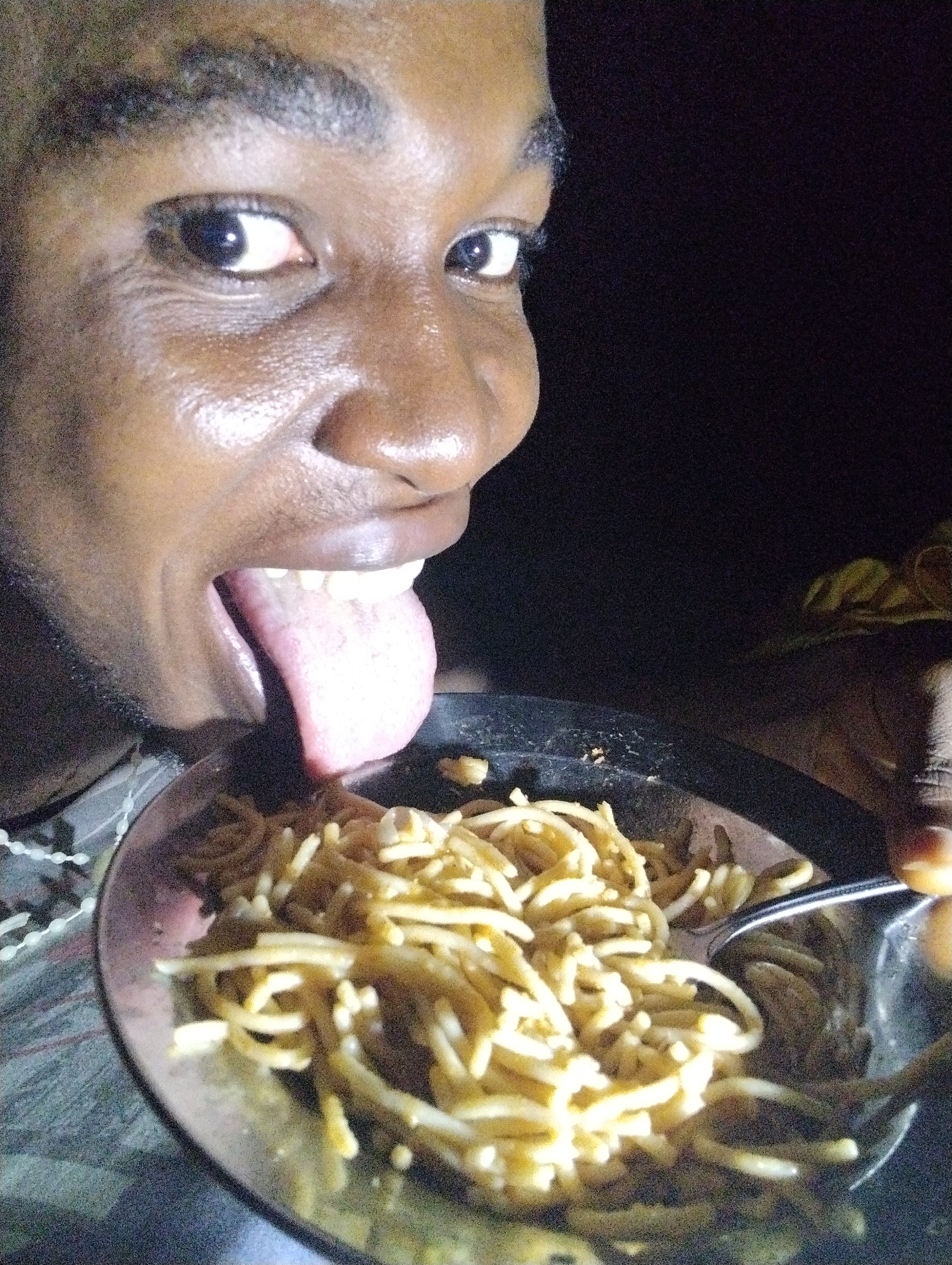 The image shows a close-up of a person, likely a young adult male with dark skin, playfully engaging with a bowl of food. The person's face is visible from the eyes to the mouth, with their left eye wide open and looking towards the food. Their tongue is extended outwards and downwards, towards a metallic bowl filled with light-colored noodles or spaghetti. This action suggests a humorous, eager, or teasing interaction with the meal.
A fork, presumably silver, is partially submerged in the noodles within the bowl on the right side. The background is very dark, indicating that the photo was taken at night or in a dimly lit indoor setting. The person's face and the food are brightly illuminated, likely by a camera flash or a direct light source. Their skin appears somewhat shiny. On the far left, a white, possibly beaded or patterned item is faintly visible. There is no visible text in the image. The scene captures a lighthearted moment during a meal, potentially in Ihiagwa, Nigeria.