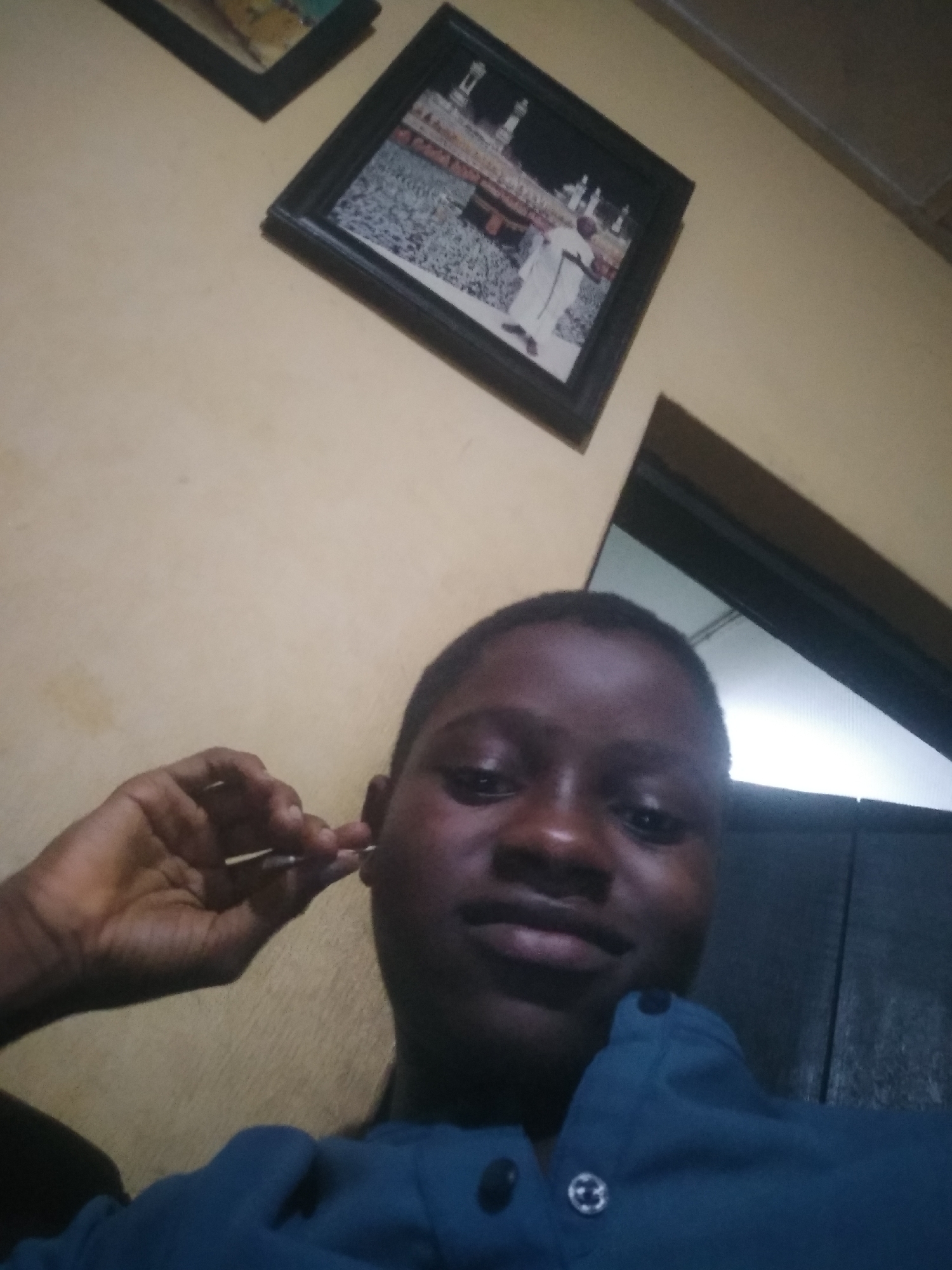 A medium-shot selfie shows a dark-skinned person holding a pair of tweezers to their ear. The person has short, dark hair and is wearing a dark blue shirt. The background is a light yellow wall with two framed pictures hanging on it. A dark doorway or partition is also visible. The location is consistent with a domestic interior. While the question prompts suggest a location in Shangisha, Nigeria, the image alone does not provide sufficient information to confirm this.