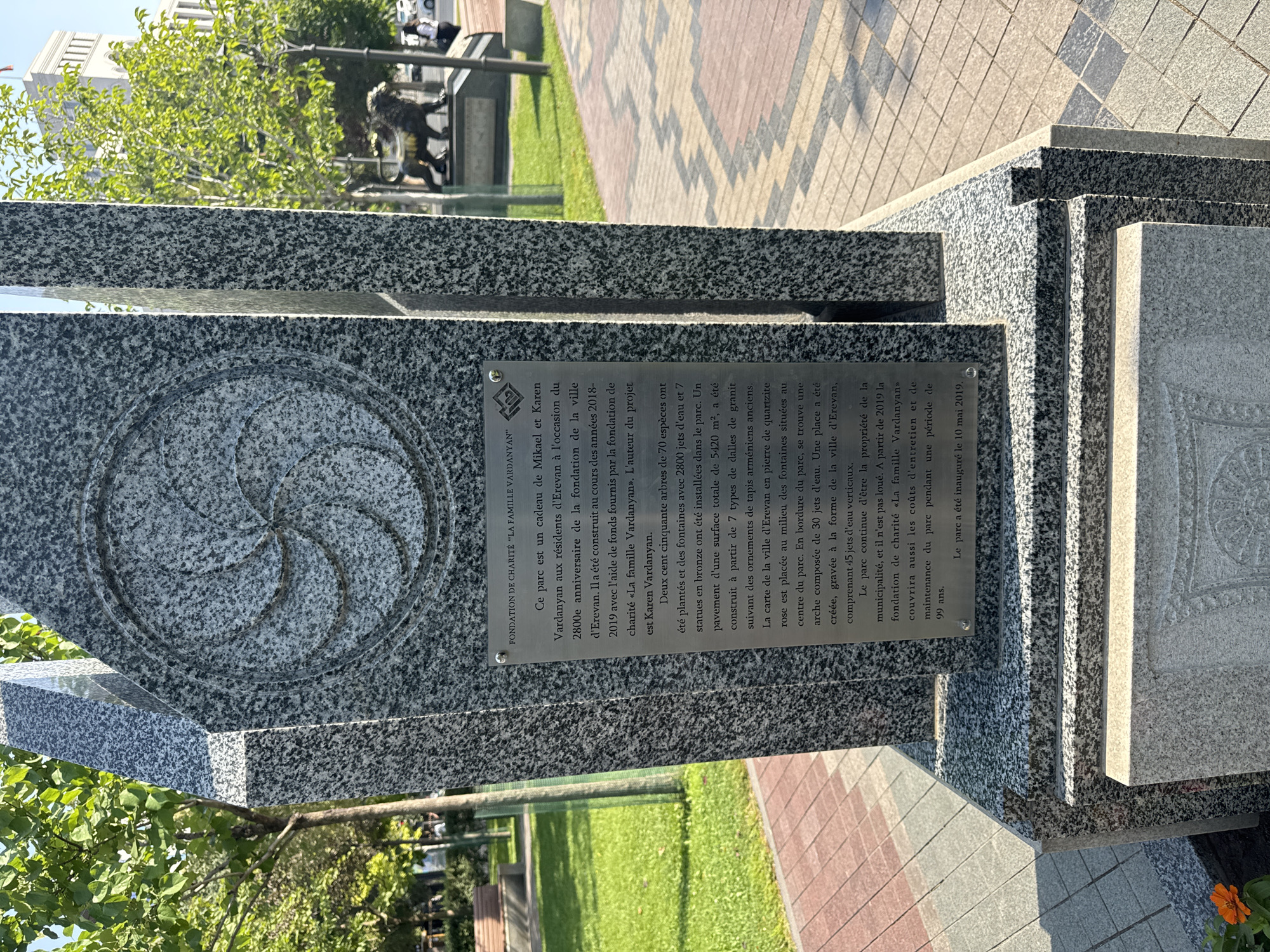 A close-up, eye-level shot captures a memorial monument in a city park, likely in Yerevan, Armenia, due to the inscription's reference to "la ville d'Erevan." The monument is constructed from polished granite, featuring a large, circular, carved Armenian eternity symbol on its front face. A silver plaque is affixed to the front of the monument, displaying text in French. Behind the monument, a section of paved walkway with a geometric pattern is visible, transitioning to a grassy area. Further in the background, a bronze lion statue is situated, and a building with a distinct architectural style stands in the distance, possibly a government building or a significant landmark. Sunlight casts shadows, indicating it is daytime, and the scene is set against a backdrop of green trees and a clear blue sky.