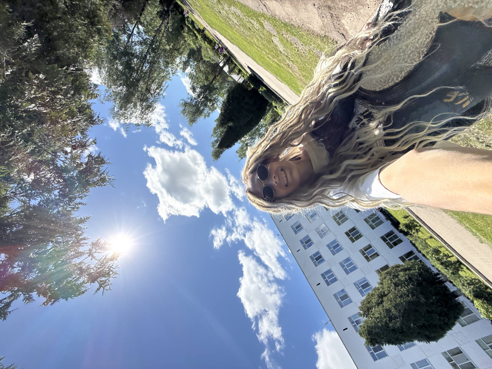 A person with long blonde hair, wearing round sunglasses, a white shirt, and a shawl stands outside. The person is positioned against a backdrop of a clear blue sky with scattered clouds and a bright sun obscured by a tree's foliage in the bottom left corner. In the background, there is a white building with multiple windows and green vegetation. The photo appears to be taken from a low angle, looking up at the subject. While Rusanovo, Russia is specified in the request, the image does not contain sufficient information to confirm the location.