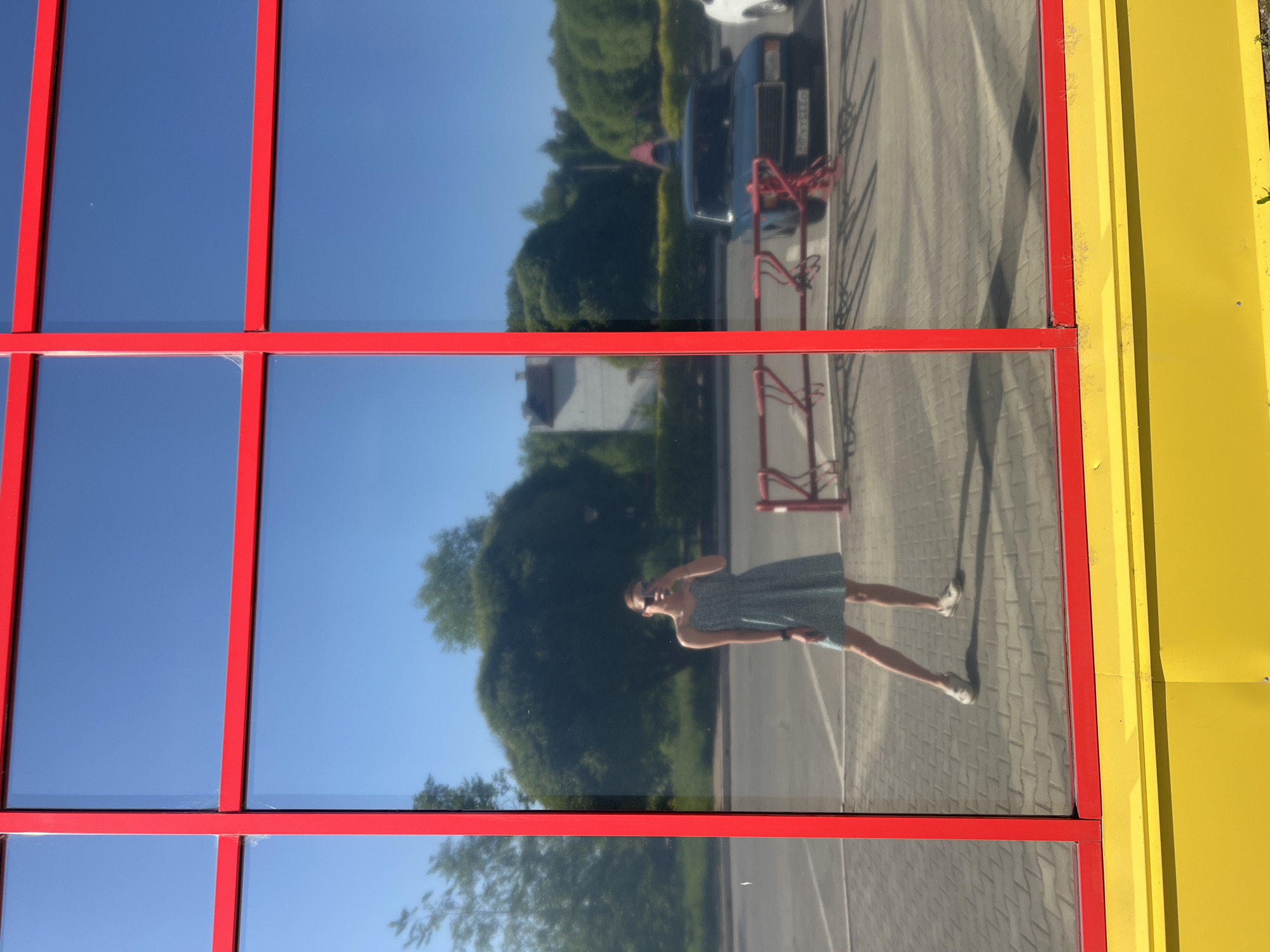 The image depicts the reflection in a building's windows in the city of Chaikovsky, Russia. The windows are framed in red, and the building has yellow siding on the right. The reflection shows trees, a car with a license plate partially visible, a red bicycle rack, pavement, and a person in a dress taking a photograph with a phone. The sky is clear and blue.