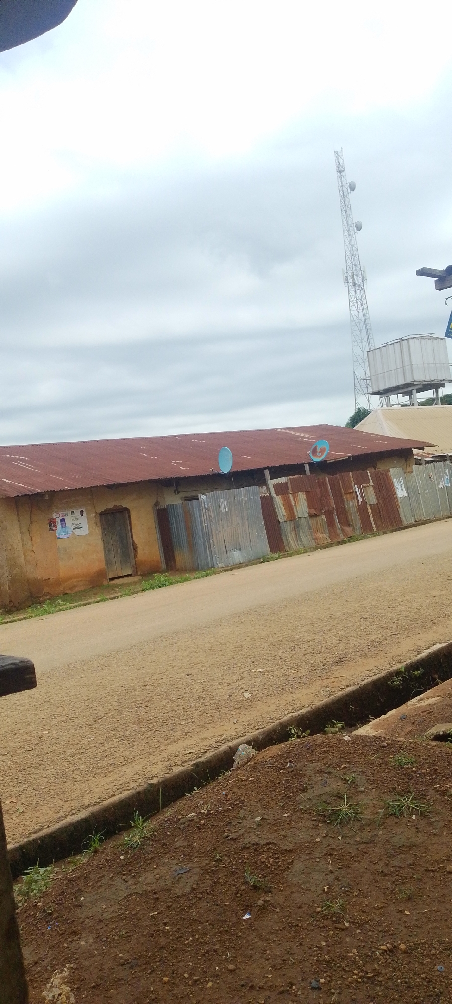 This image depicts an outdoor scene in Baissa, Nigeria. The foreground shows a dirt road with a curb and some patchy vegetation and soil. In the middle ground, a long, weathered building with a rusty corrugated metal roof dominates the view. The building appears to be made of a light-colored, possibly stucco or mud material, with a wooden door and some sections covered in corrugated metal. A few posters are affixed to the wall near the door, one of which features a man in traditional attire with text that appears to be related to a political campaign, possibly for the year 2023. Two blue satellite dishes are mounted on the roof.
In the background, a tall, lattice-like telecommunications tower stands prominently against an overcast sky. To the right of the tower, a large water tank is visible, situated on a raised platform. The weather appears to be cloudy, suggesting it might be an overcast day or evening. The overall impression is one of a rural or semi-urban environment in Nigeria, with elements of infrastructure like communication towers and water storage alongside older, possibly more rudimentary buildings. No specific activity or event is clearly depicted, and the scene appears to be a still, environmental shot.