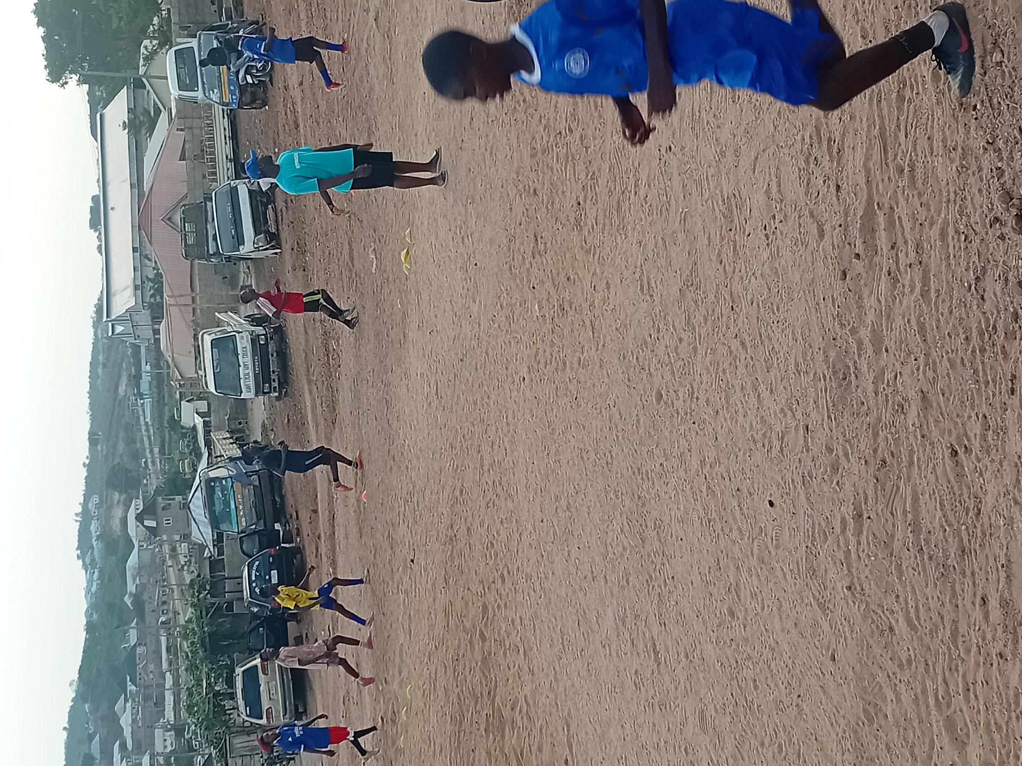 The image depicts an outdoor scene, likely a training ground or open dirt area, with several individuals engaged in physical activity. The primary subjects are approximately ten people, mostly young males, dressed in athletic wear, including soccer jerseys and shorts. They are spread across a wide, unpaved expanse of light brown sand or dirt, which serves as the foreground and midground. Some individuals are actively in motion, appearing to run or stretch, while others are standing. Small yellow cones or markers are visible on the ground near some of the participants, suggesting organized drills or training.
In the background, a variety of parked vehicles are visible, including several trucks, vans, and cars, lining what appears to be a dirt road or parking area adjacent to the training space. One white truck prominently displays "SAM LOCAL GVT TRUCK" and "TOYOTA" on its side. Behind the vehicles, a collection of buildings, including houses and a larger structure with a pitched roof, can be seen, indicating a semi-urban or rural settlement. Distant hills or mountains rise on the horizon under a bright, possibly overcast, sky.
The time of day appears to be daytime, with soft, diffused lighting suggesting either an overcast sky or perhaps early morning/late afternoon. The overall atmosphere is one of focused activity or sports practice. A person in a blue soccer jersey and dark shorts is prominently featured in the top right foreground, captured mid-stride. Another individual in a light blue t-shirt and dark shorts stands further back, near one of the parked trucks. The ground is uneven, with visible tire tracks and foot impressions in the sand.