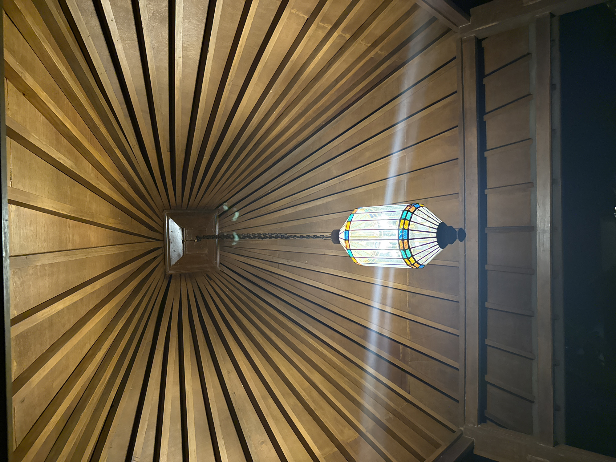 The image captures a unique architectural ceiling structure, likely indoors or within a covered outdoor space in Kuta Selatan, Indonesia. The primary subject is a wooden ceiling composed of numerous parallel planks. These planks are arranged radially, converging dramatically towards a central, recessed square opening, creating a striking sense of depth and perspective.
A decorative lamp, resembling a stained-glass lantern with multi-colored panels (blue, yellow, green, orange), hangs from this central point by a dark chain. The lamp is illuminated, casting a warm glow and producing bright streaks of light that reflect off the wooden surfaces, emphasizing the plank lines. The wood itself is a warm, light brown tone, with natural grain visible, and some planks appear darker in the recesses, adding to the visual texture.
To the right side of the frame, a segment of a darker wooden wall or column is visible, featuring horizontal paneling. The area beyond this structure is dark, suggesting either an unlit interior space or the nighttime outdoors. The overall ambiance is warm and inviting, highlighted by the intricate wooden craftsmanship and the decorative light fixture. No people or visible text are present in the image.