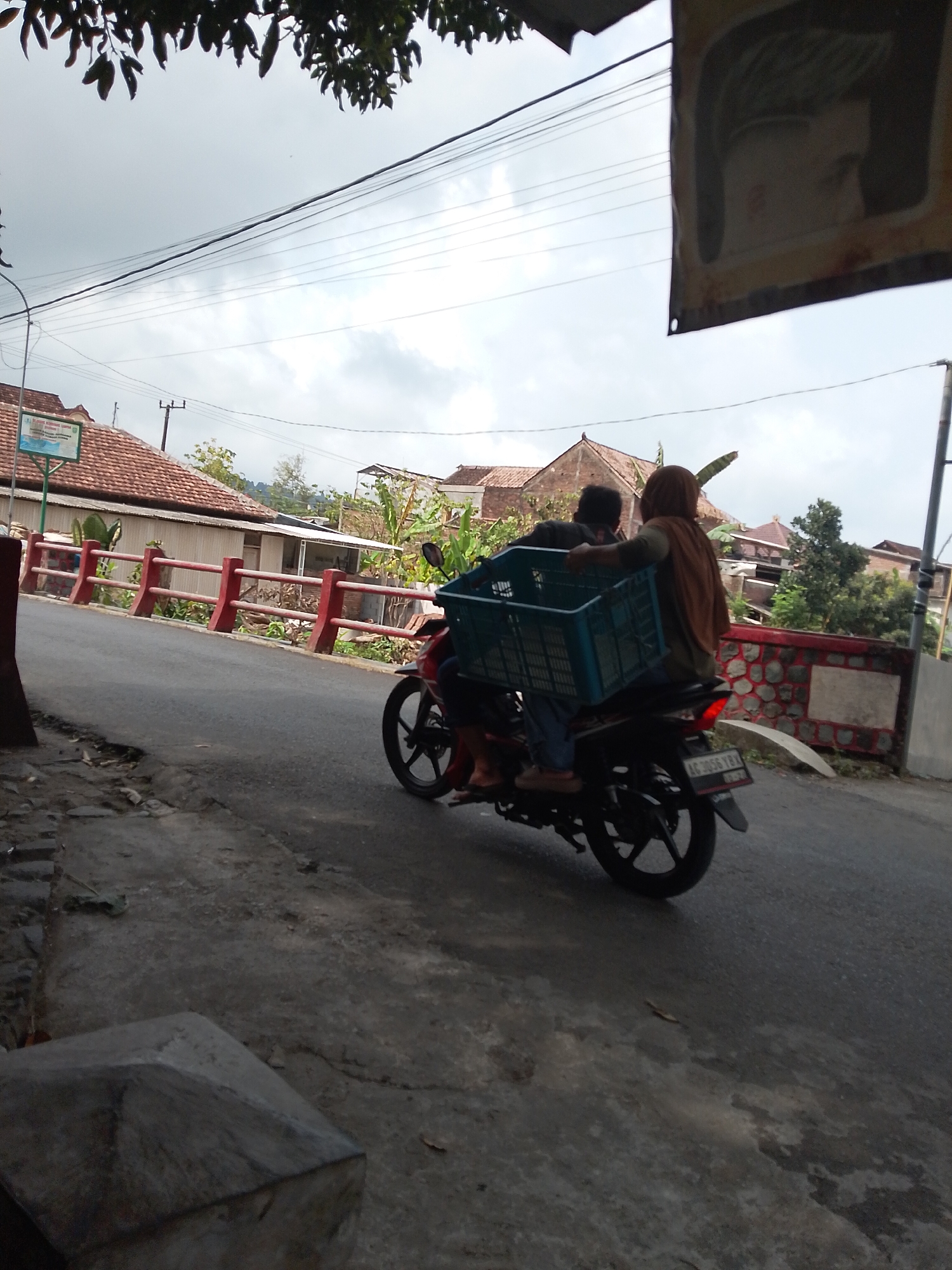 The image depicts a street scene in Tasikmadu, Indonesia, likely during daylight hours under an overcast sky. The focal point is a motorcycle traveling on the road. Two individuals are on the motorcycle; the driver is in front and appears to be a male wearing dark clothing. Behind the driver, a female wearing a brown headscarf and a brown garment is seated. They are transporting a large, teal-colored plastic crate loaded with items. The crate is positioned between them, extending over the back of the motorcycle. The motorcycle has a visible license plate that reads "AG 3056 TBX".
The environment includes a paved road with a slightly uneven surface, suggesting a somewhat rural or less maintained urban area. To the left of the road, a rough, textured embankment with some debris is visible. On the right side of the road, a red and white fence with vertical posts separates the road from an area with buildings and vegetation. The buildings have terracotta-tiled roofs, characteristic of Indonesian architecture. In the background, more houses and trees are discernible. Overhead, power lines stretch across the frame, and a portion of a banner or sign is visible in the upper right corner, partially obscuring the sky. The overall impression is a snapshot of daily life and transportation in the specified location.