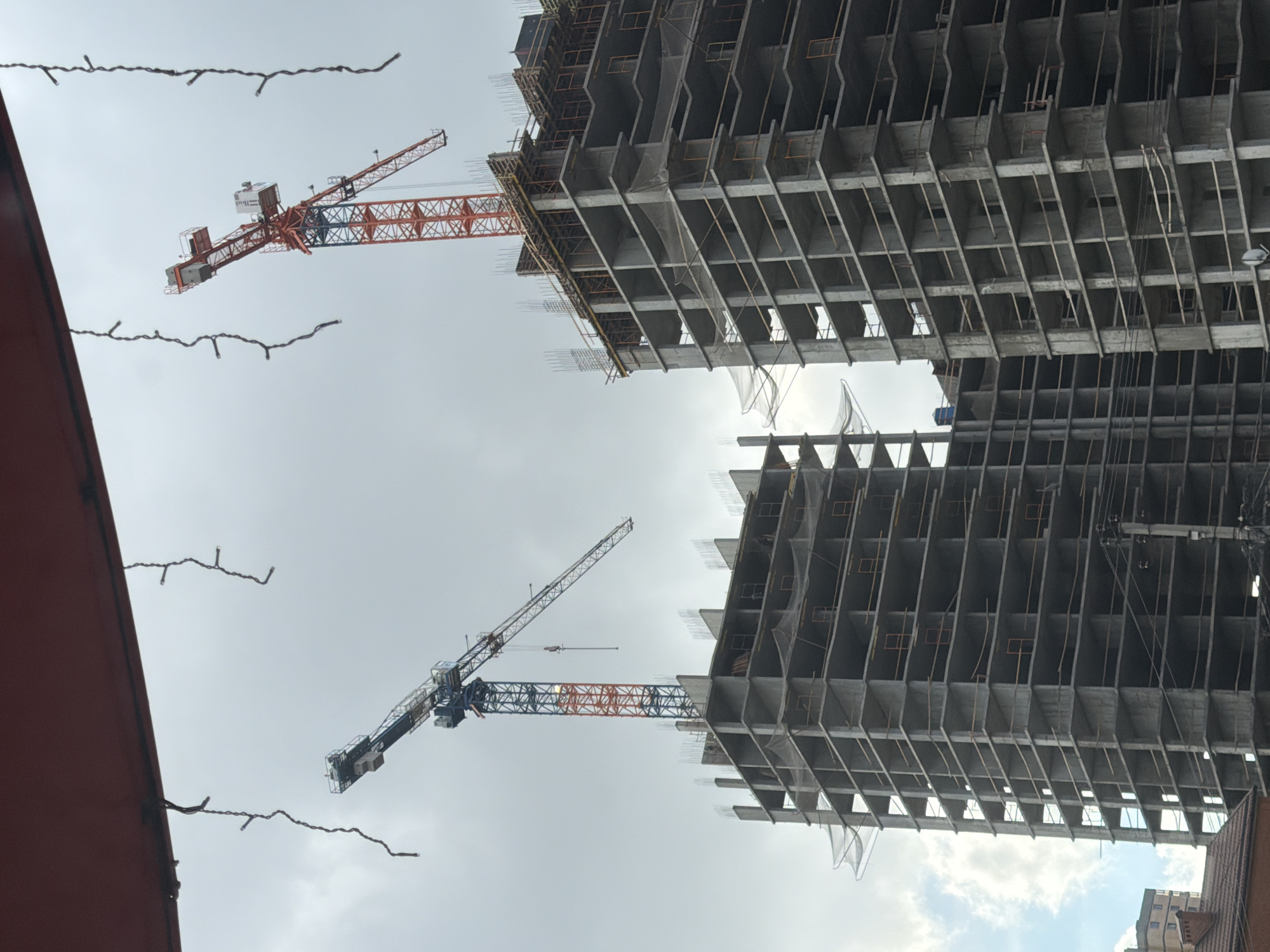 The image shows two unfinished high-rise buildings under construction in what is likely Rostov-on-Don, Russia, given the prompt. Two construction cranes, one predominantly orange and the other blue and orange, stand adjacent to the buildings. The structures are multi-story concrete skeletons with exposed rebar. A portion of a building roof is visible in the lower right corner of the picture, and strands of string lights dangle from the upper left. The sky is overcast with patches of blue visible near the horizon.