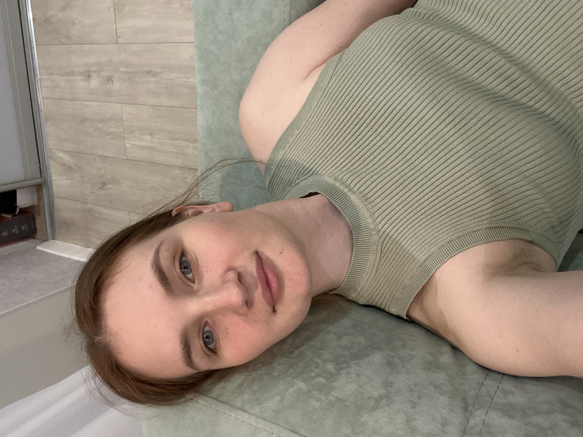 A young woman with fair skin, light brown hair, and blue eyes is the primary subject of this close-up, head-and-shoulders shot. She is lying down, positioned diagonally in the frame, and looking directly into the camera with a neutral, calm expression. A small mole or mark is visible near her mouth.
She is wearing a light olive-green, ribbed, sleeveless top. Her hair is casually parted and falls around her face and shoulders. Her left arm is visible, bent at the elbow, and shows some visible underarm hair.
The woman is resting on a soft, padded surface, likely a couch or bed, which is a muted sage green color, matching the general cool tone of the image.
The background on the left features a wall covered with light-colored, rectangular tiles arranged in a horizontal pattern, resembling wood grain. To the far left, a vertical metallic or white frame, possibly part of a mirror, door, or shower enclosure, is visible. Below the woman's head, a hint of a white sheet or blanket can be seen. The setting appears to be an indoor space in Chaikovsky, Russia, possibly a modern bathroom, bedroom, or a treatment room, characterized by clean lines and subdued colors. The lighting suggests daytime. There is no visible text in the image.