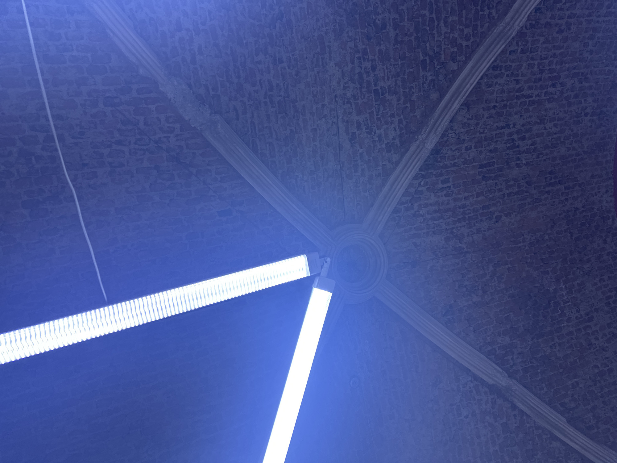 The image shows the interior of a building, possibly located in Saint Petersburg, Russia. The perspective is from below, looking up at a brick vaulted ceiling, reinforced with white or light-colored plaster ribs that intersect at a central point, possibly a decorative medallion. Two bright fluorescent lights are positioned on the left side of the frame, pointing towards the viewer. A thin white wire is visible extending from the upper left corner. The scene is illuminated with a strong blue light, likely from the fluorescent fixtures. The overall impression is one of a historical structure with modern lighting.