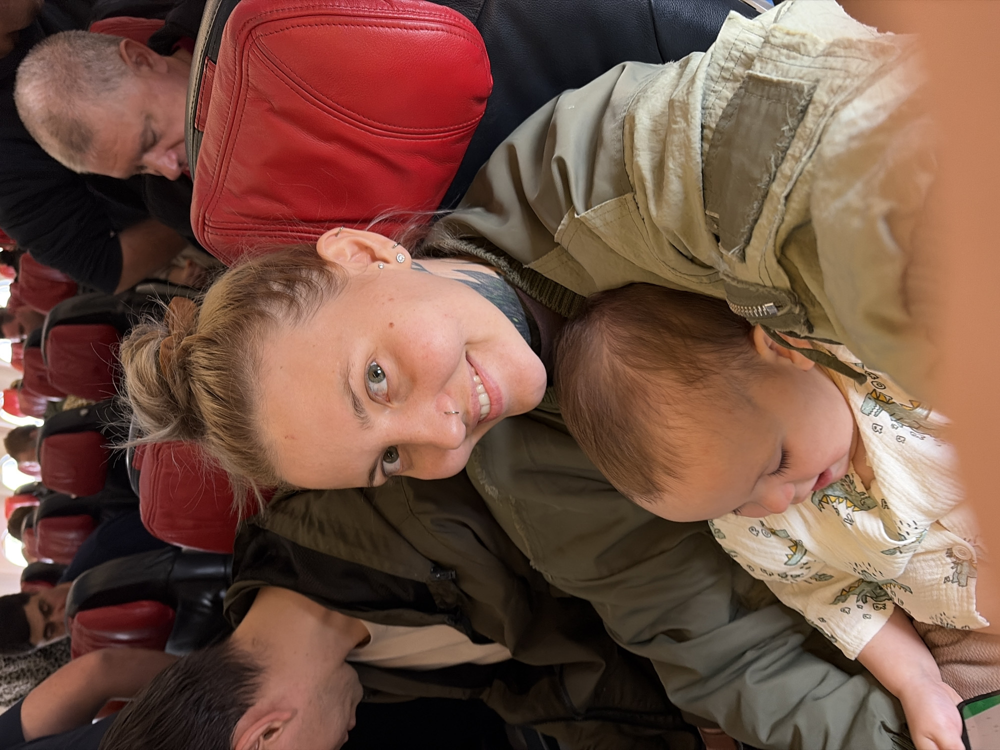 The image shows a woman with short blonde hair pulled back in a bun, wearing an olive green jacket and a white top, smiling and holding a baby in her arms. The baby, wearing a white button-down shirt with dinosaur prints, appears to be sleeping. The woman has multiple ear piercings and a neck tattoo.
In the background, there are other people seated in what seems to be an airplane cabin, judging by the red headrests and arrangement of seats. A bald man is sleeping in the upper left corner. Another person is visible in the bottom left corner.
The location in the city of Khimki, Russia is not determinable from the image.