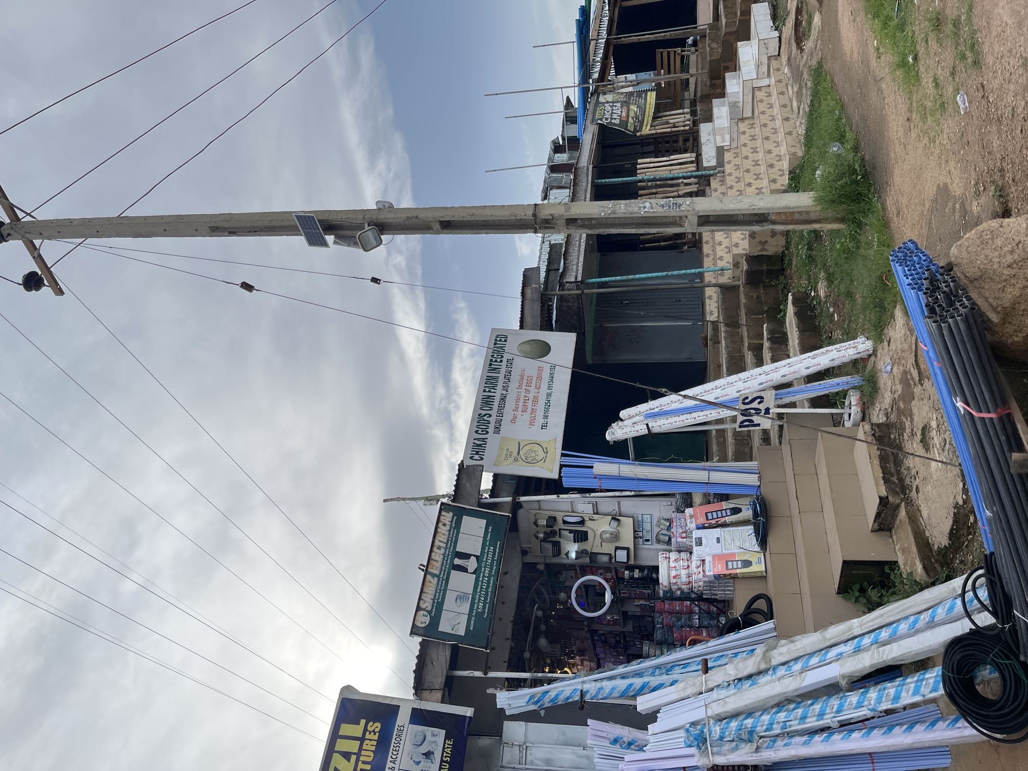 This media file depicts a street view in Gura Top, Nigeria. The scene is dominated by a utility pole with power lines crisscrossing the sky, suggesting an overcast or hazy day. Attached to the pole are a solar panel and a light fixture. Below the pole, a row of shops or stalls is visible, displaying various goods for sale.
The shops appear to specialize in electrical supplies and building materials. Signs for "SAMZ ELECTRICAL" and "CHIKA GOD'S OWN FARM INTEGRATED" are prominently displayed. The electrical store showcases an array of light fixtures and wiring, while the "CHIKA GOD'S OWN FARM" sign indicates services related to egg supply and poultry feeds and accessories. Piles of blue and grey pipes, likely for construction or plumbing, are stacked on the ground in front of the shops.
In the foreground, more building materials, including white and blue-striped pipes or conduits, are neatly arranged. The ground is a mix of dirt and grass, with some scattered litter. The overall impression is of a busy commercial area in a Nigerian town, with an emphasis on construction and agricultural supplies. The presence of the solar panel on the utility pole hints at efforts towards alternative energy solutions in the area. Visible text includes business names, addresses, and contact information.